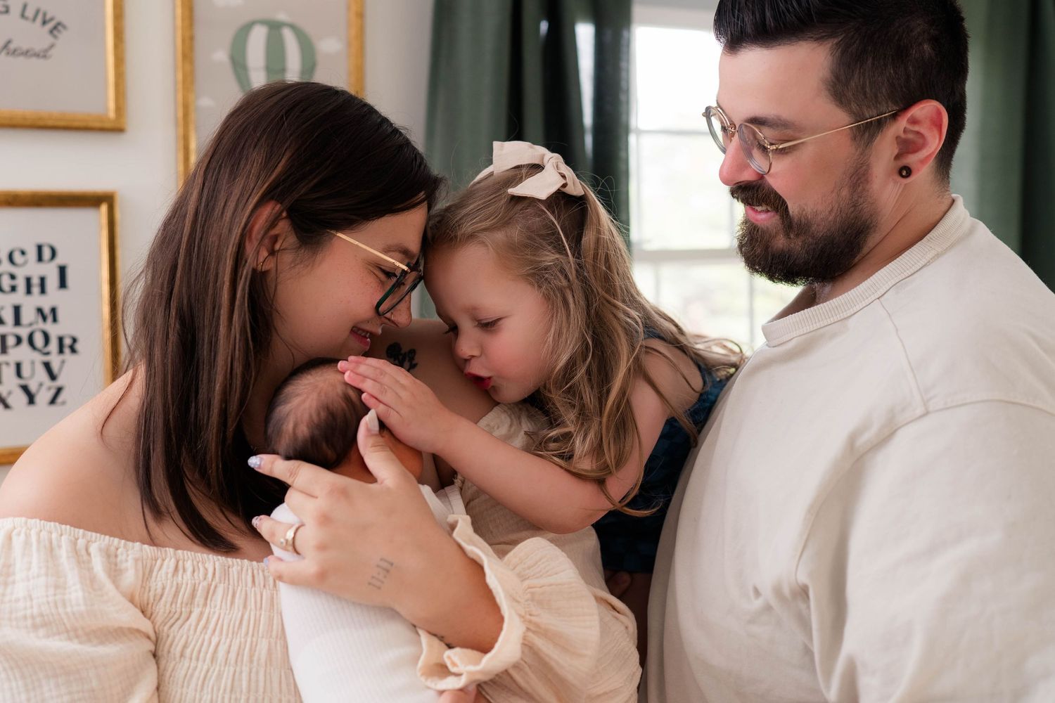 A young family shares a tender moment, with parents and toddler girl lovingly gazing at a newborn baby indoors.