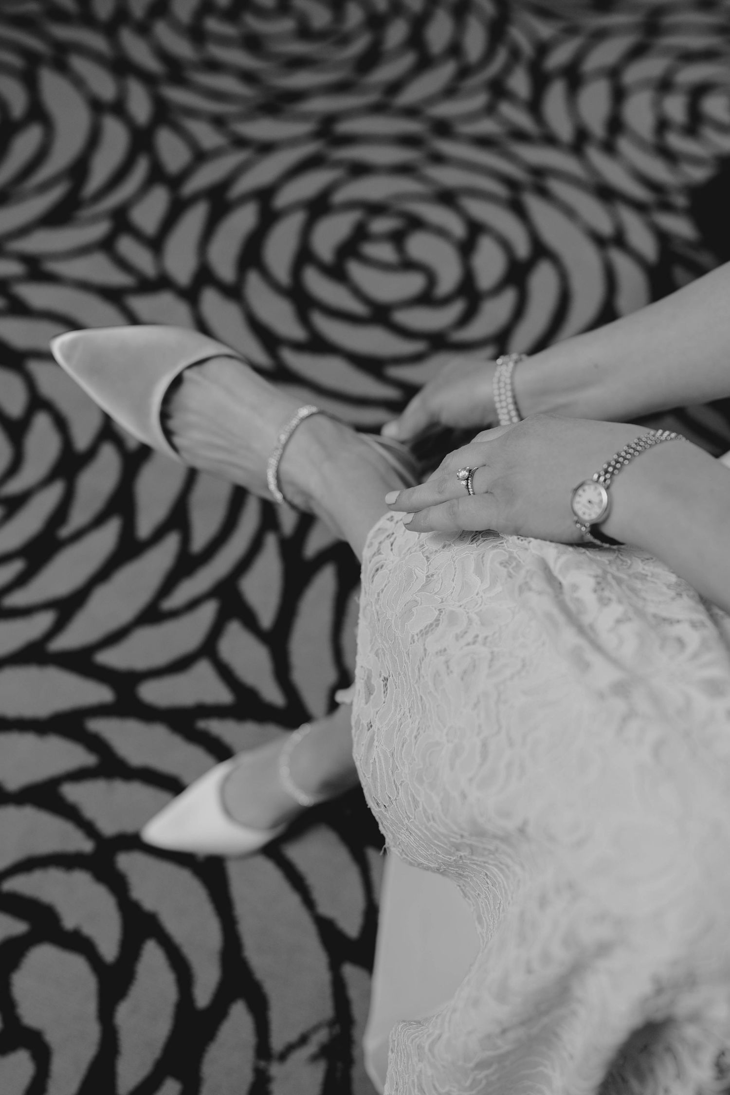 Black and white photo of a bride adjusting her white heels, wearing a watch and bracelet on her wrist.