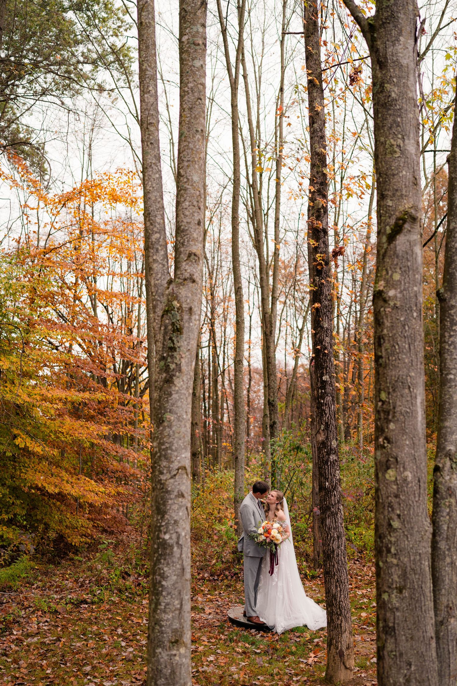 Bride and groom share a romantic kiss surrounded by colorful autumn trees in a forest setting.