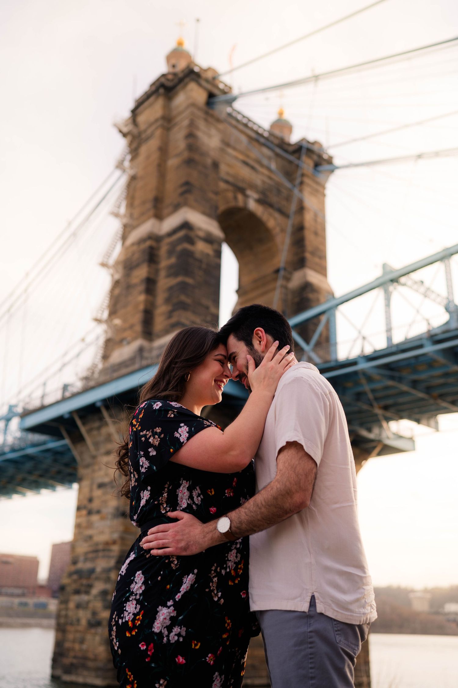 Romantic couple nearly kissing in front of a historic suspension bridge at sunset.