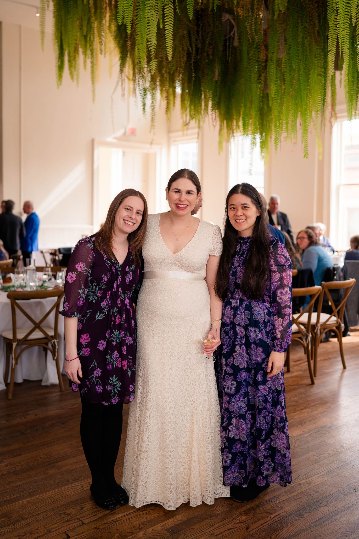 Bride in white lace gown posing with two guests under hanging ferns at wedding.