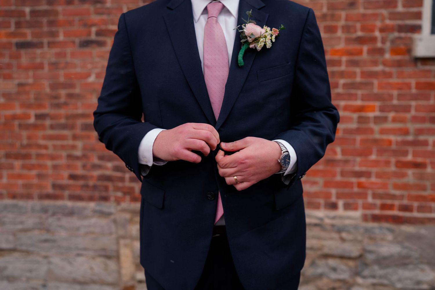 Groom in navy suit with pink tie and floral boutonniere buttoning jacket against brick wall.