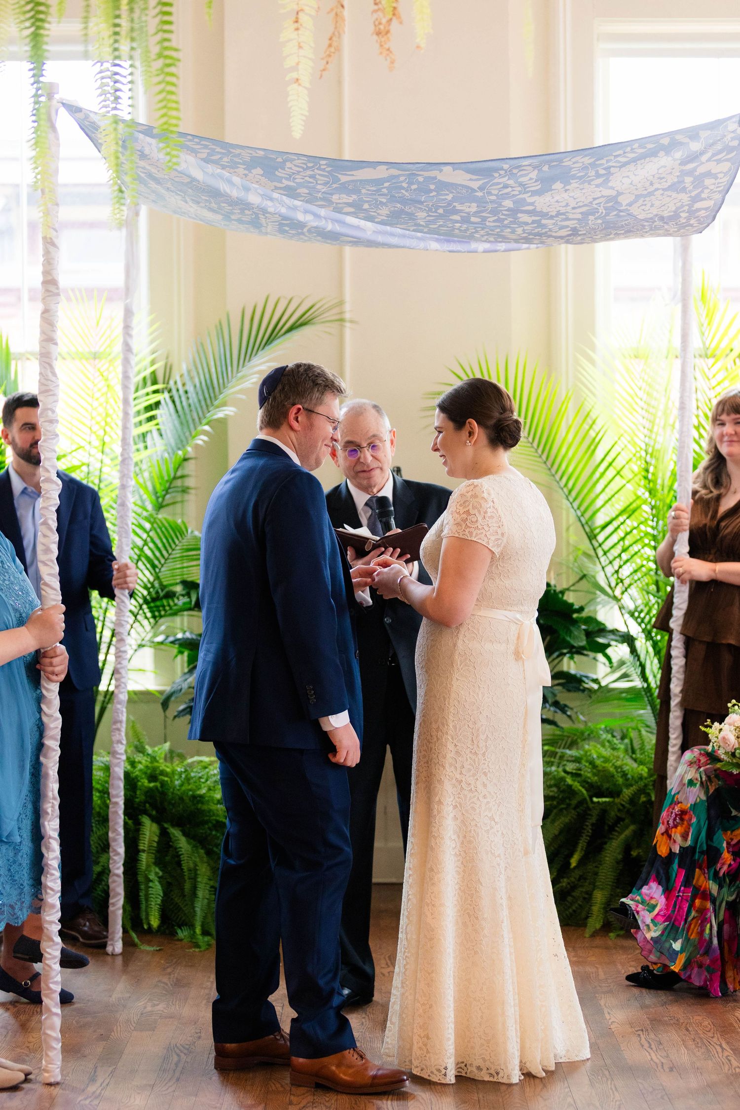 Couple holds hands under blue chuppah with officiant during Jewish wedding ceremony.