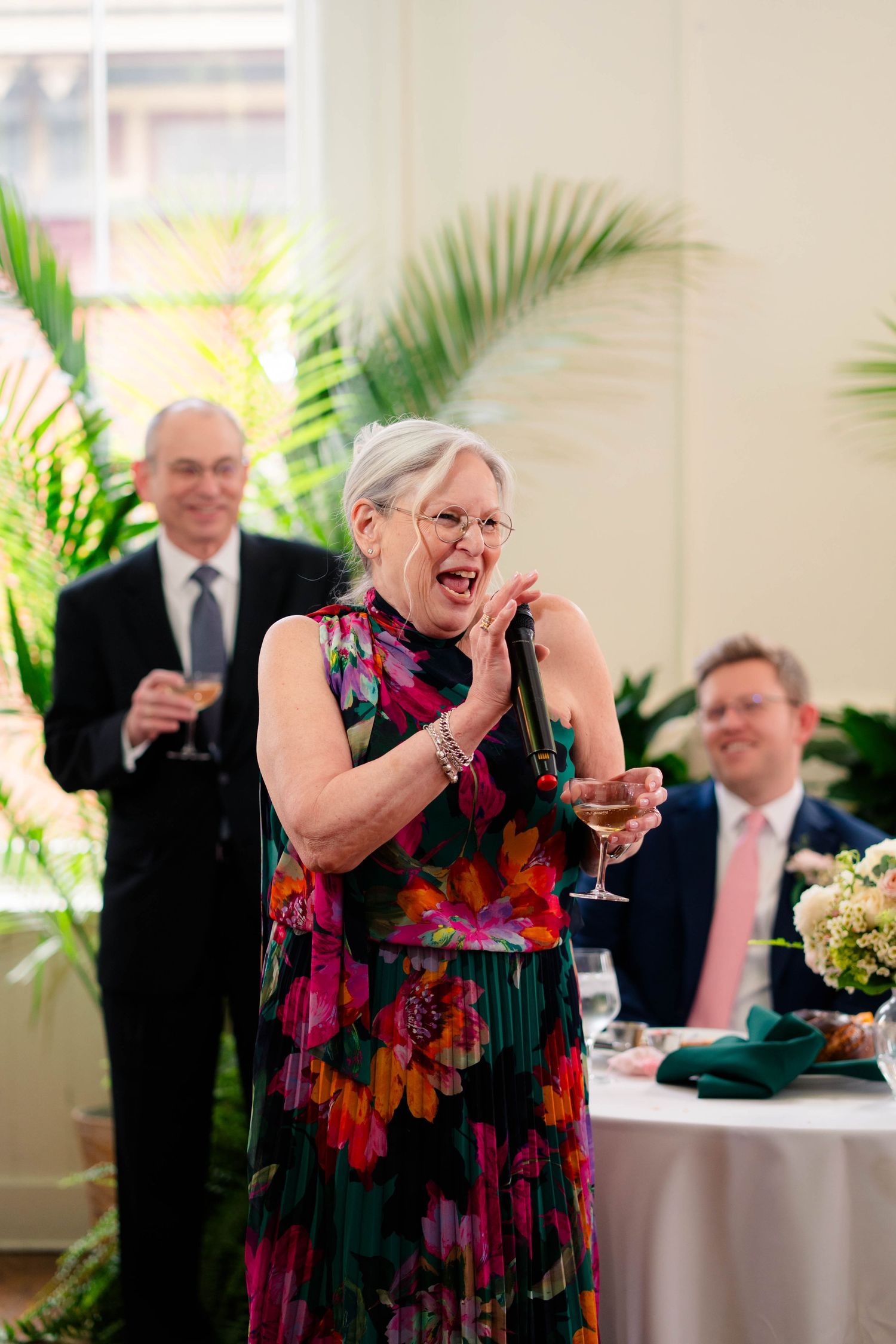 Joyful woman in vibrant floral dress delivers heartfelt wedding toast with microphone.