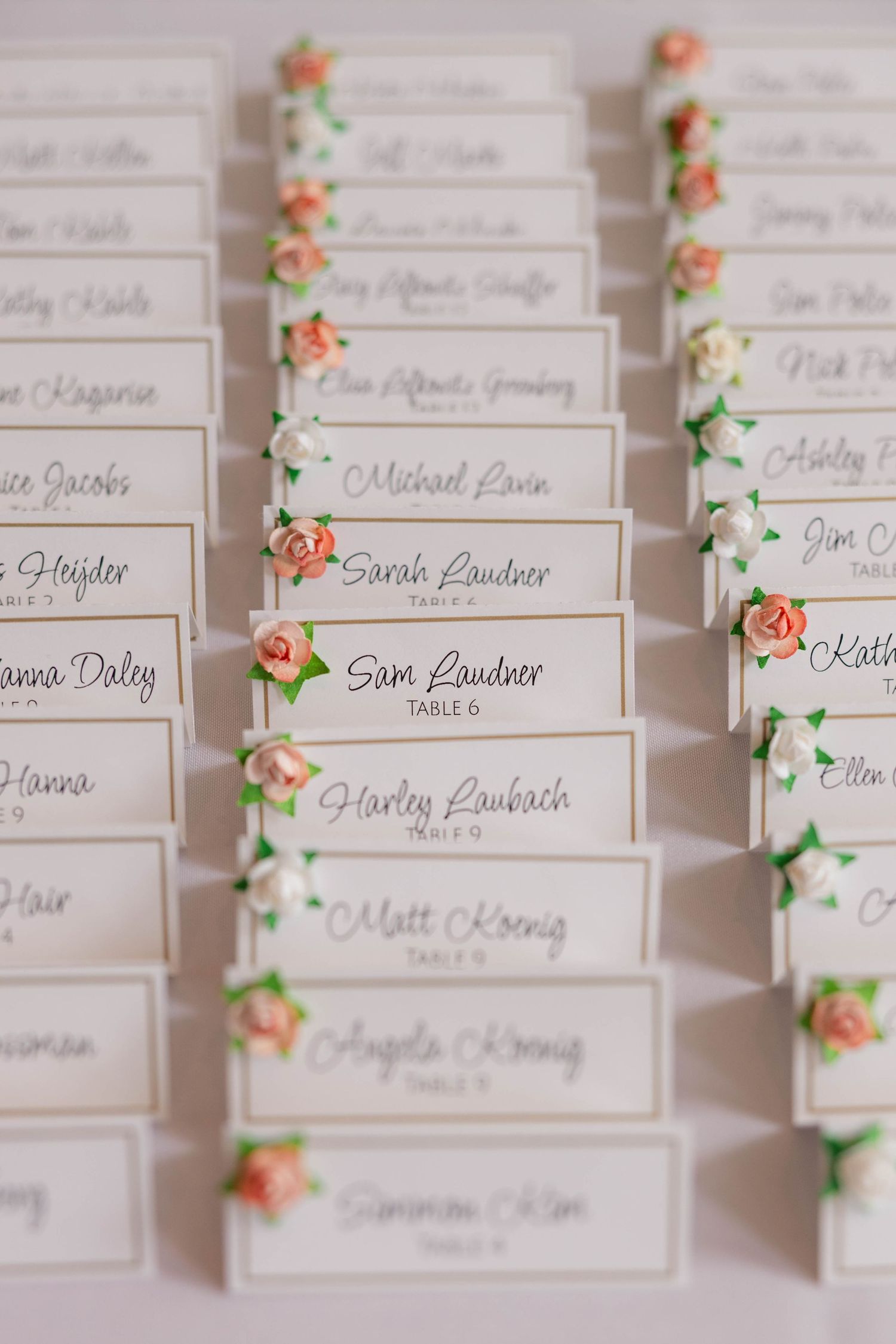 Wedding escort cards with pink rose decorations arranged in rows, featuring guest names and table assignments in calligraphy.