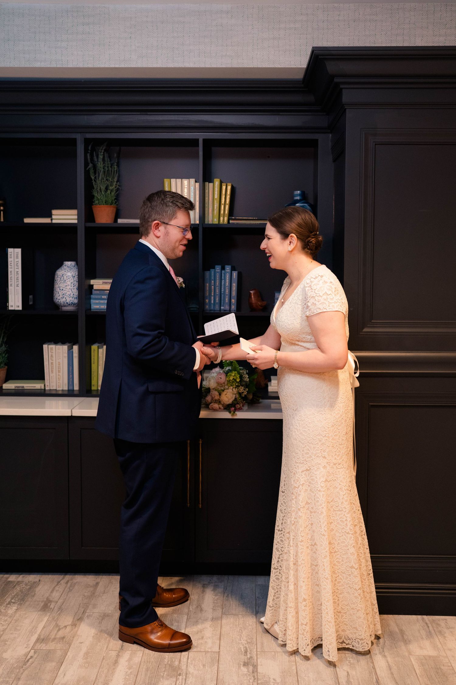 Bride and groom hold hands and smile while exchanging personal written vows in a hotel library.