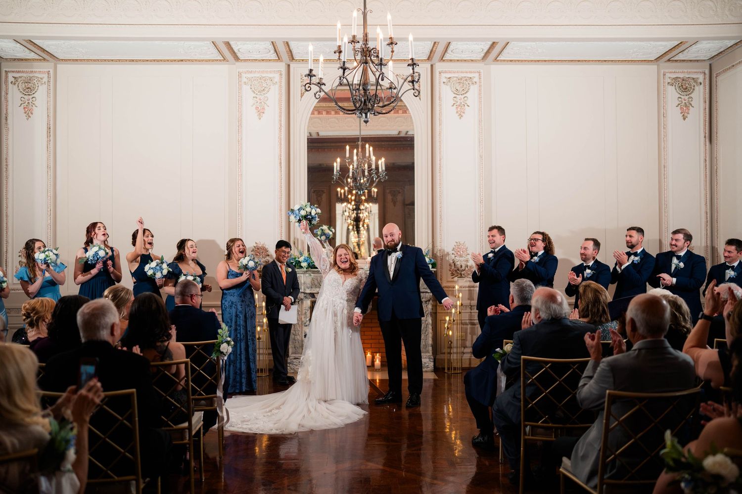 Joyful bride and groom recessional through applauding guests in ornate chandelier-lit wedding venue.