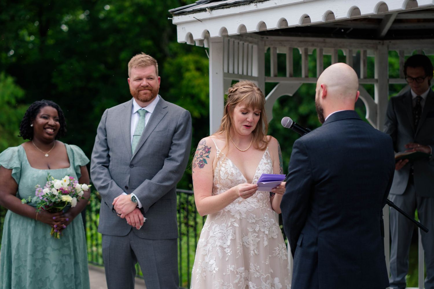 Bride reads wedding vows while groom smiles during outdoor ceremony near white gazebo with wedding party.