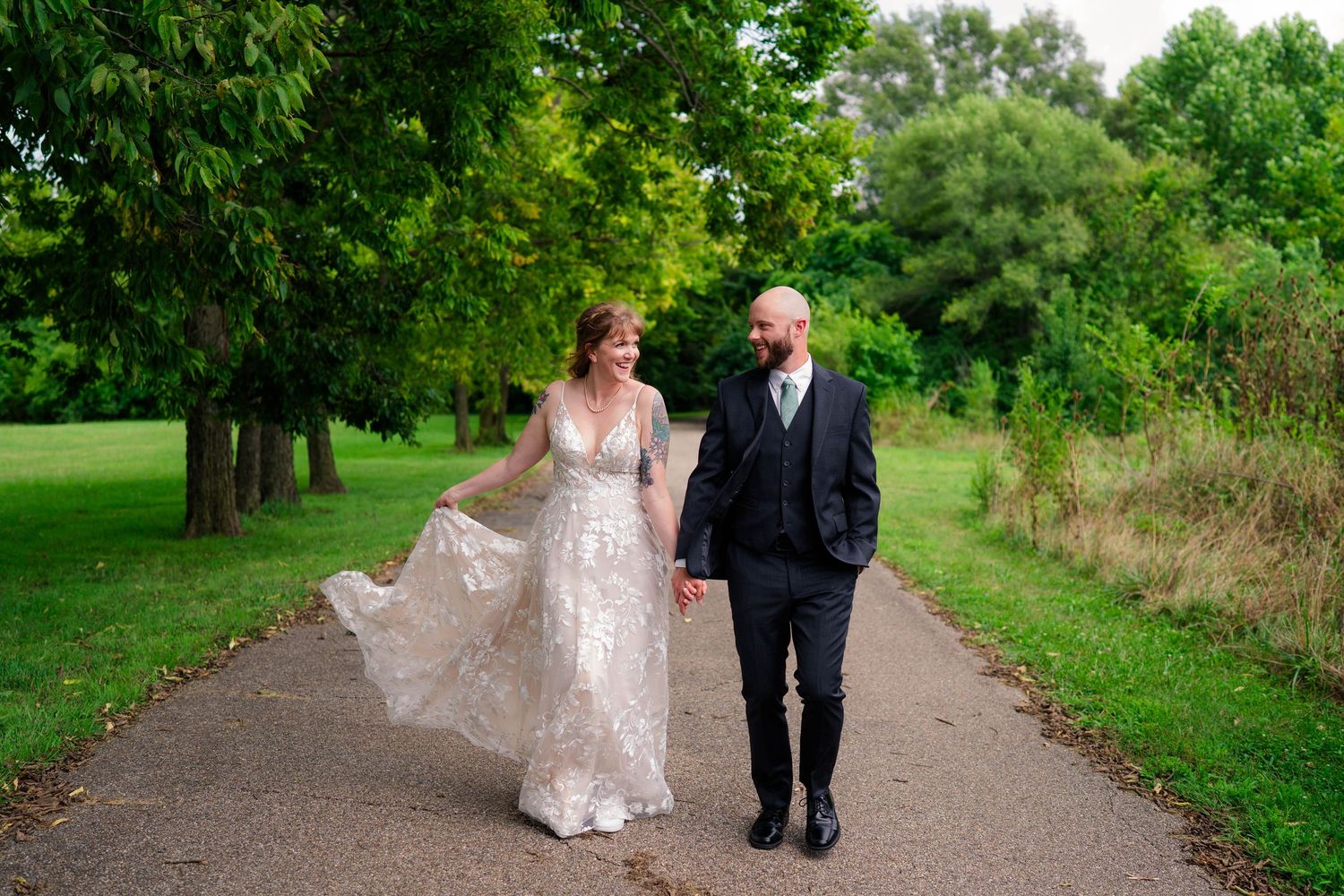 Bride in flowing lace gown and bald groom walk hand in hand along tree-lined park path.