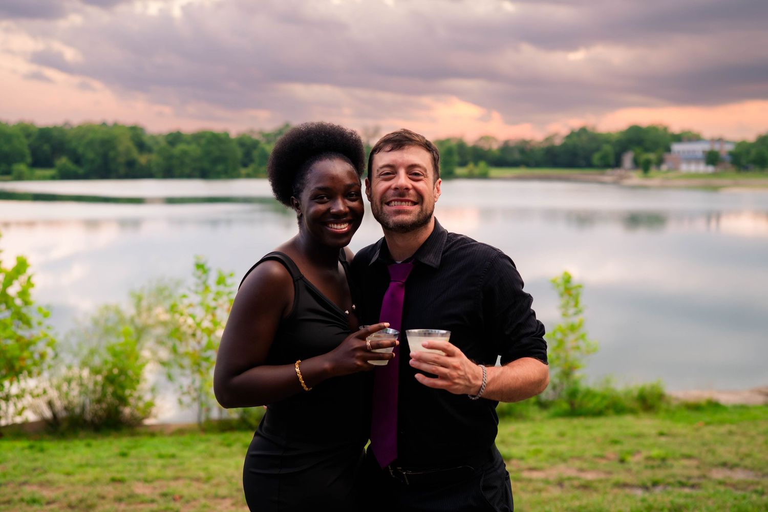 Smiling couple holds drinks while posing together near a lake at sunset during wedding.