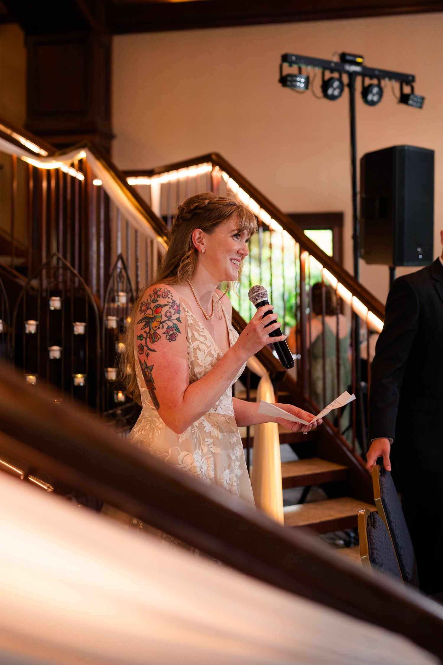 Smiling bride in lace gown holds microphone giving speech on staircase at wedding reception.