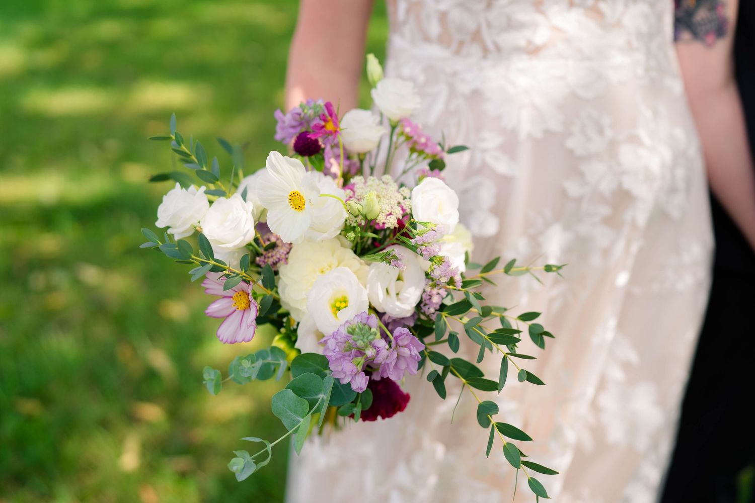 Bride holds elegant bouquet of white peonies, purple flowers, and greenery against lace wedding gown.