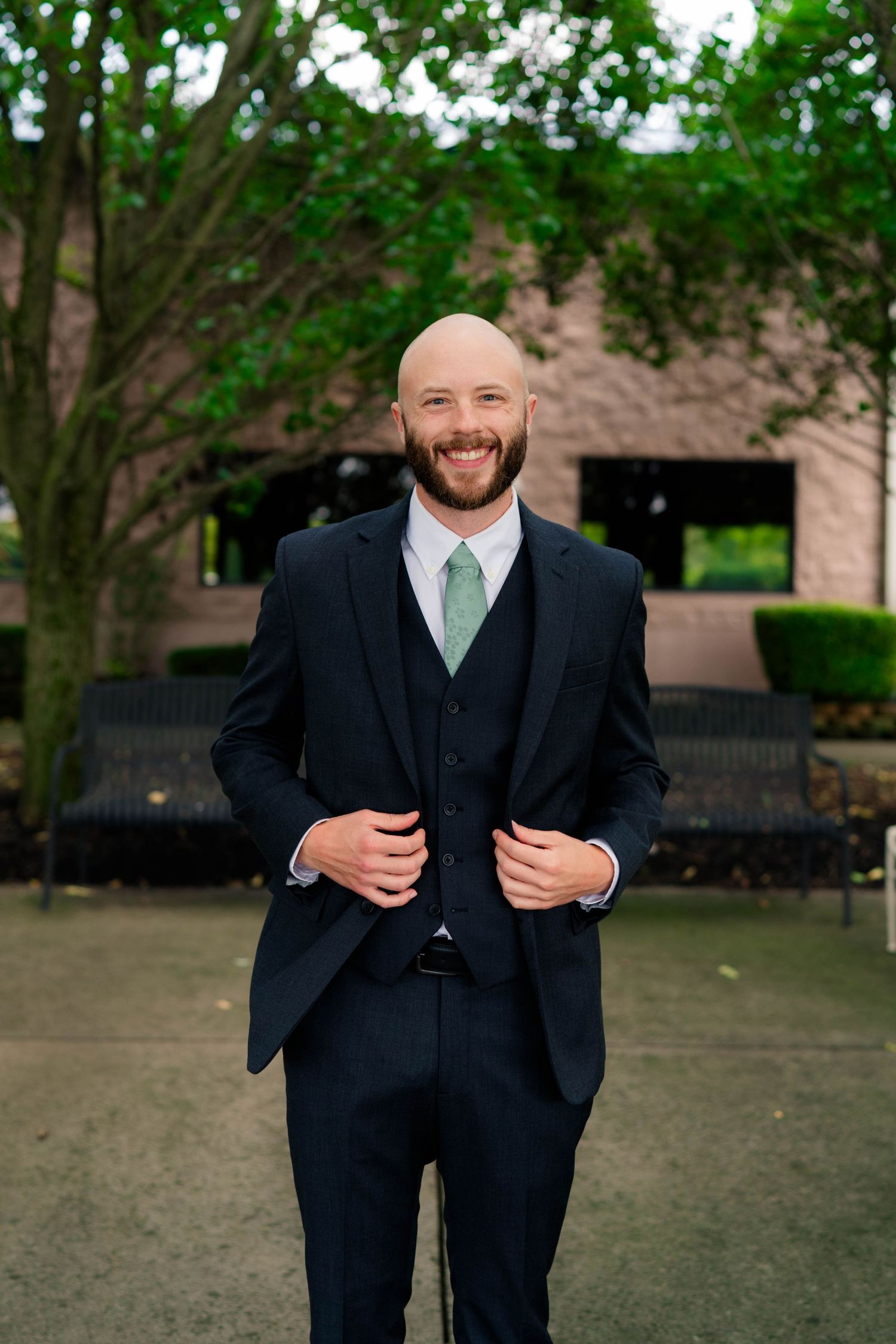Smiling bald groom in navy three-piece suit with mint green tie posing outdoors near green trees.