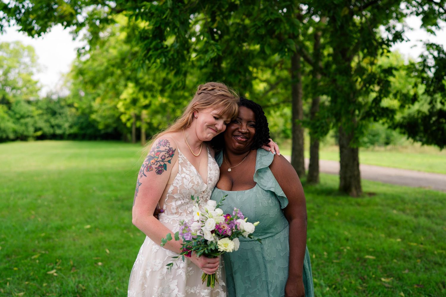 Bride with tattoos holding bouquet embracing bridesmaid in teal dress outdoors.