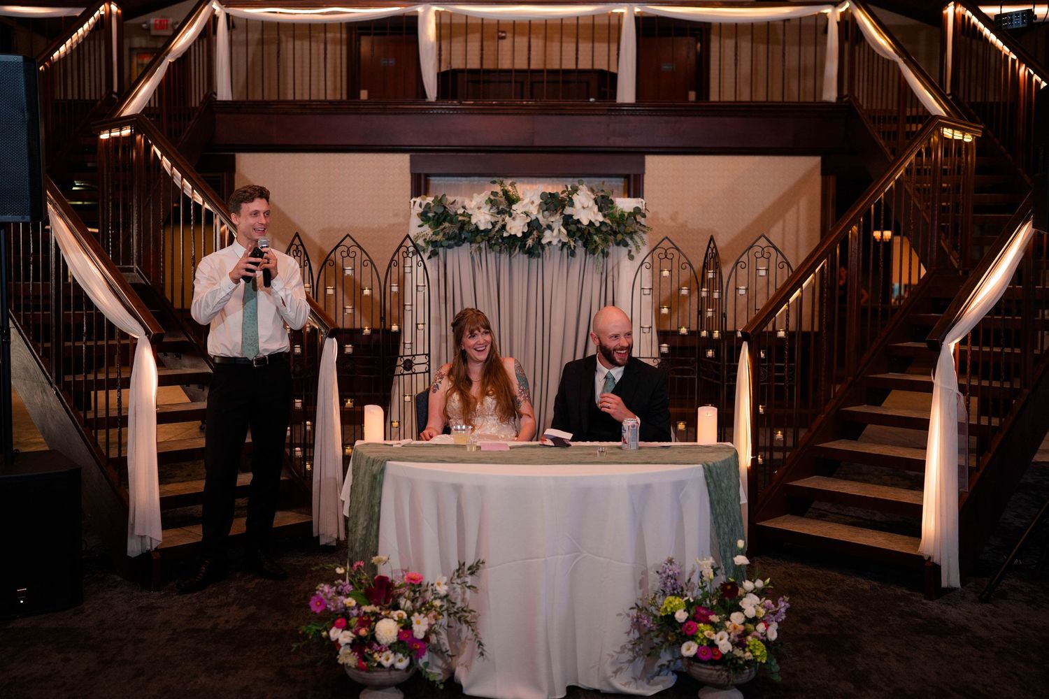 Bride and groom sign marriage documents at sweetheart table while groomsman gives toast at wedding reception.