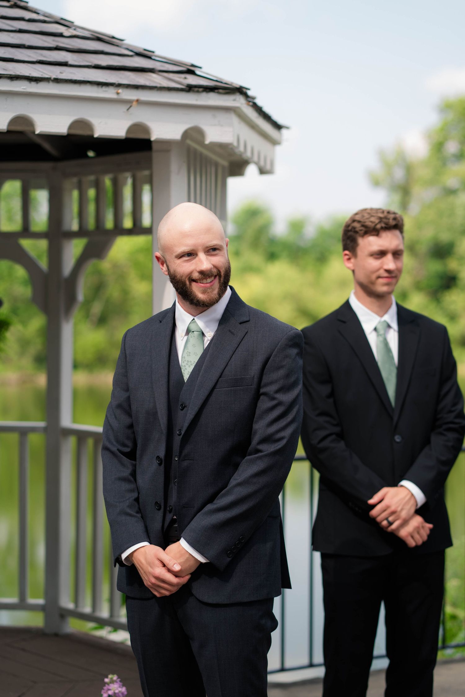 Smiling bald groom in navy suit stands at outdoor gazebo wedding ceremony.