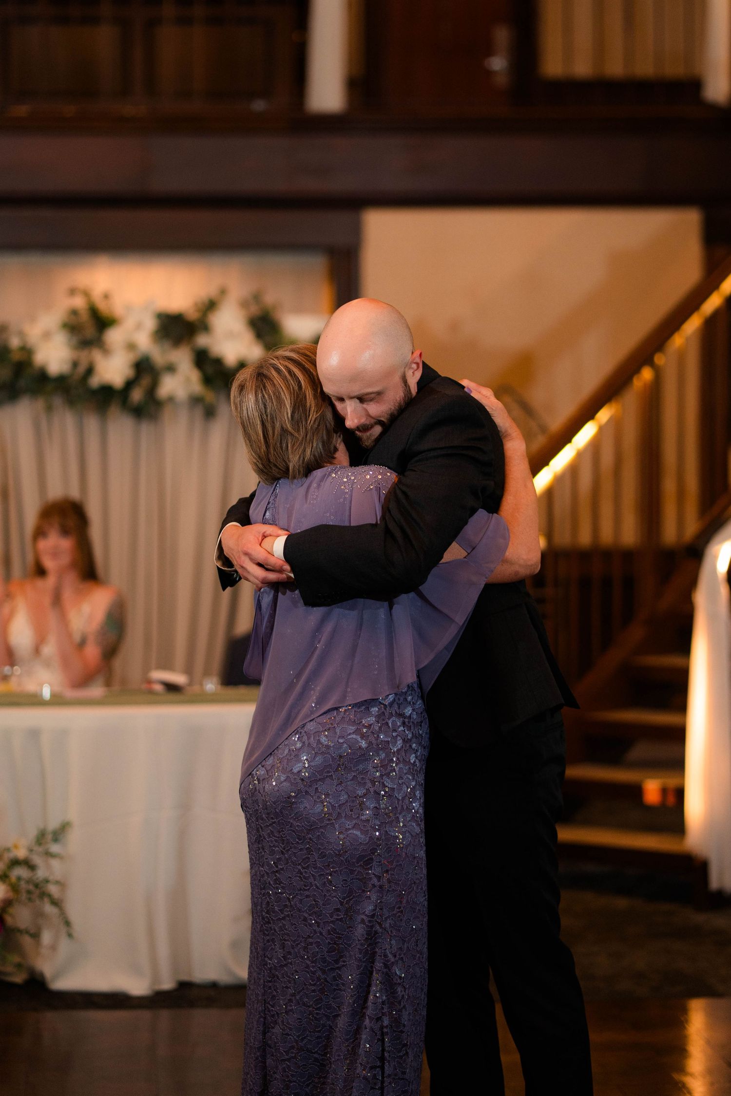 Bald groom shares emotional dance embrace with mother in purple sequin gown at wedding reception.