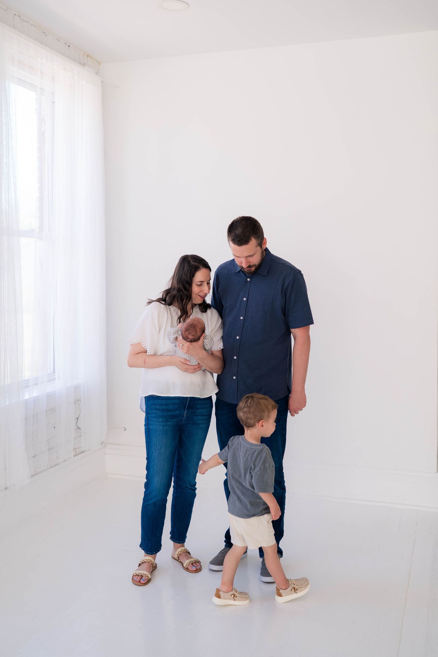 A happy family of four stands in a bright white studio, parents admiring their newborn while young son looks on curiously.