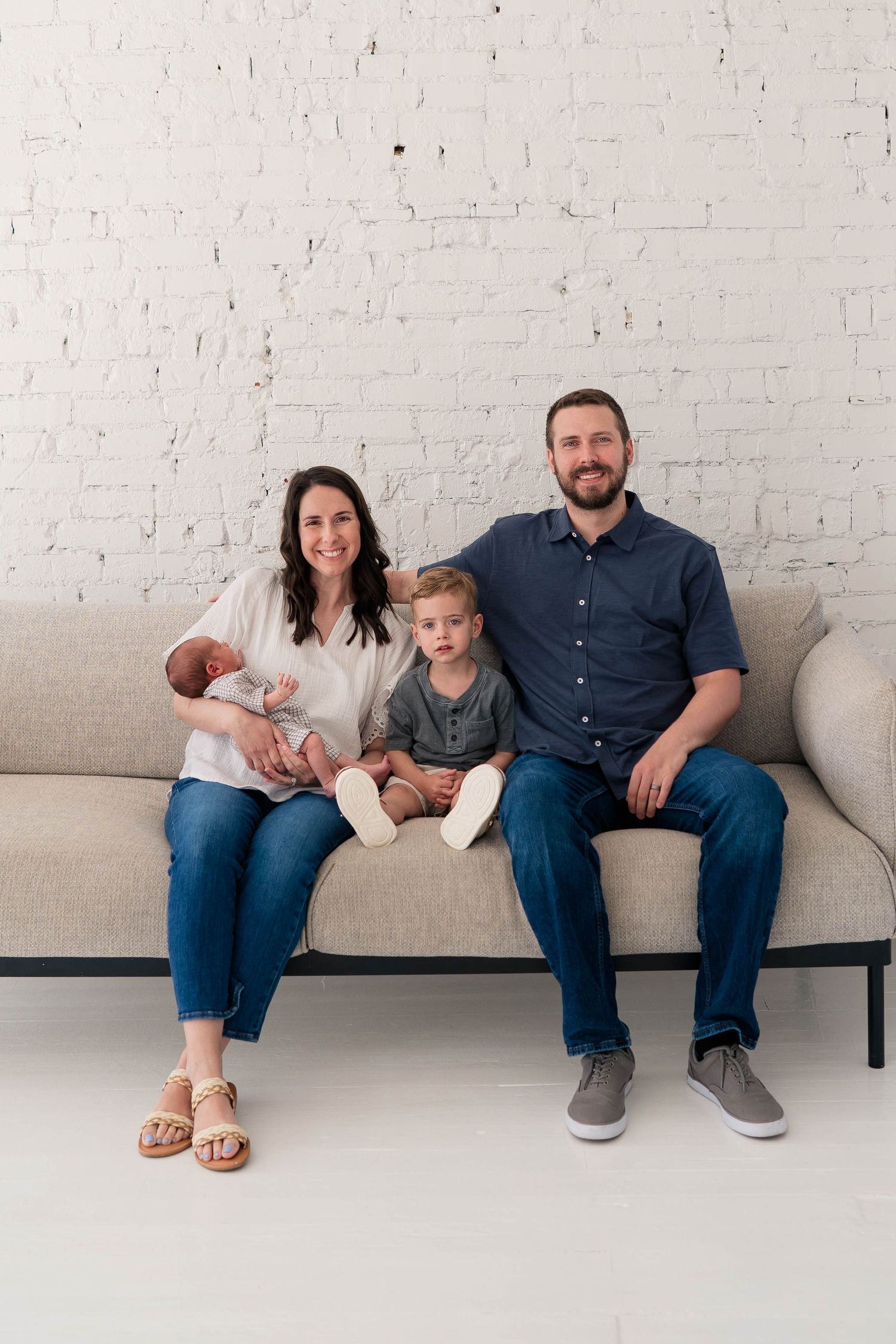 Family of four with newborn baby sitting on beige couch against white brick wall in bright studio.