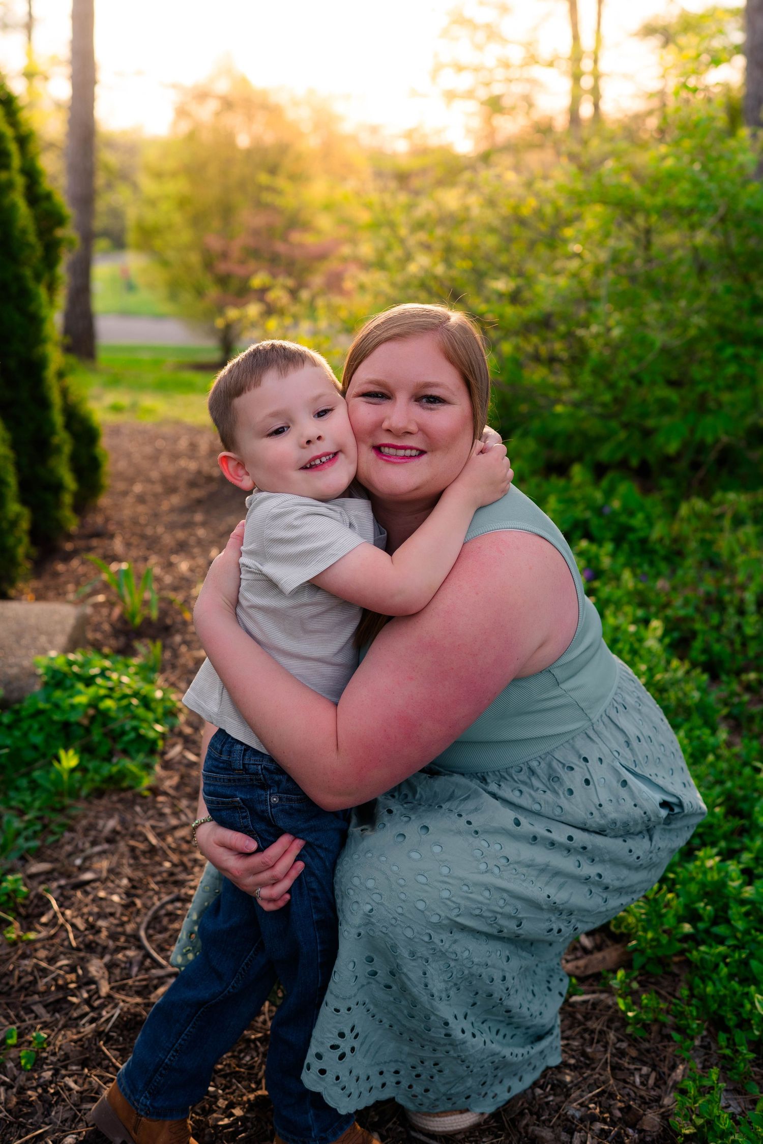 Woman in mint dress hugging young boy outdoors in garden with golden sunset light filtering through trees.
