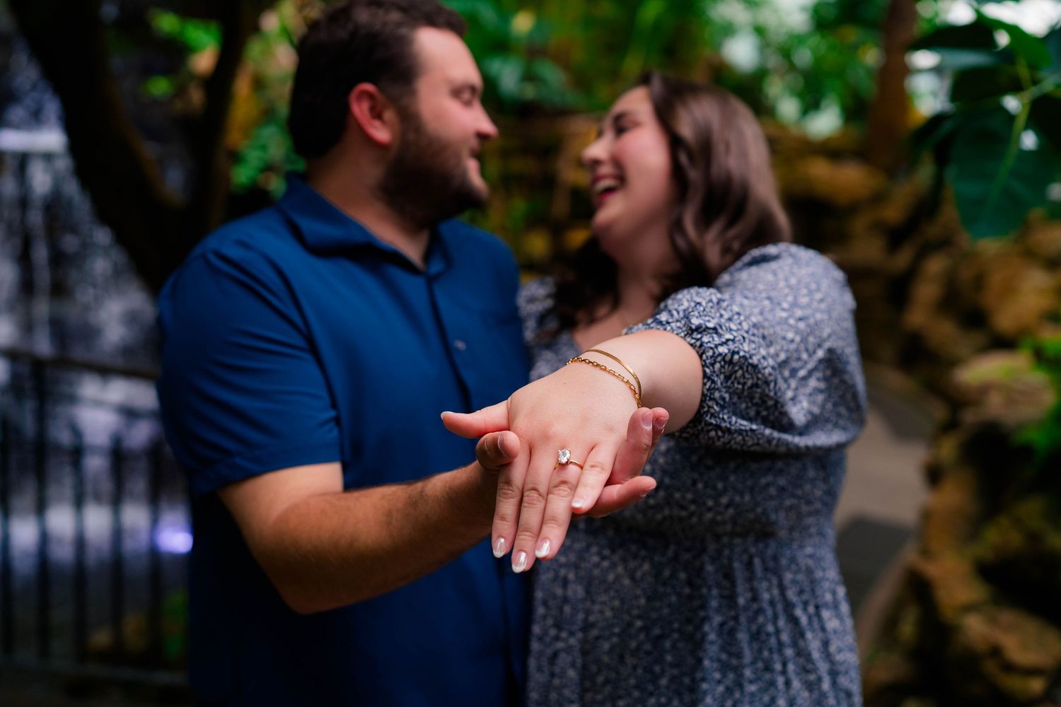 Excited newly engaged couple shows off engagement ring while laughing together in a tropical garden.