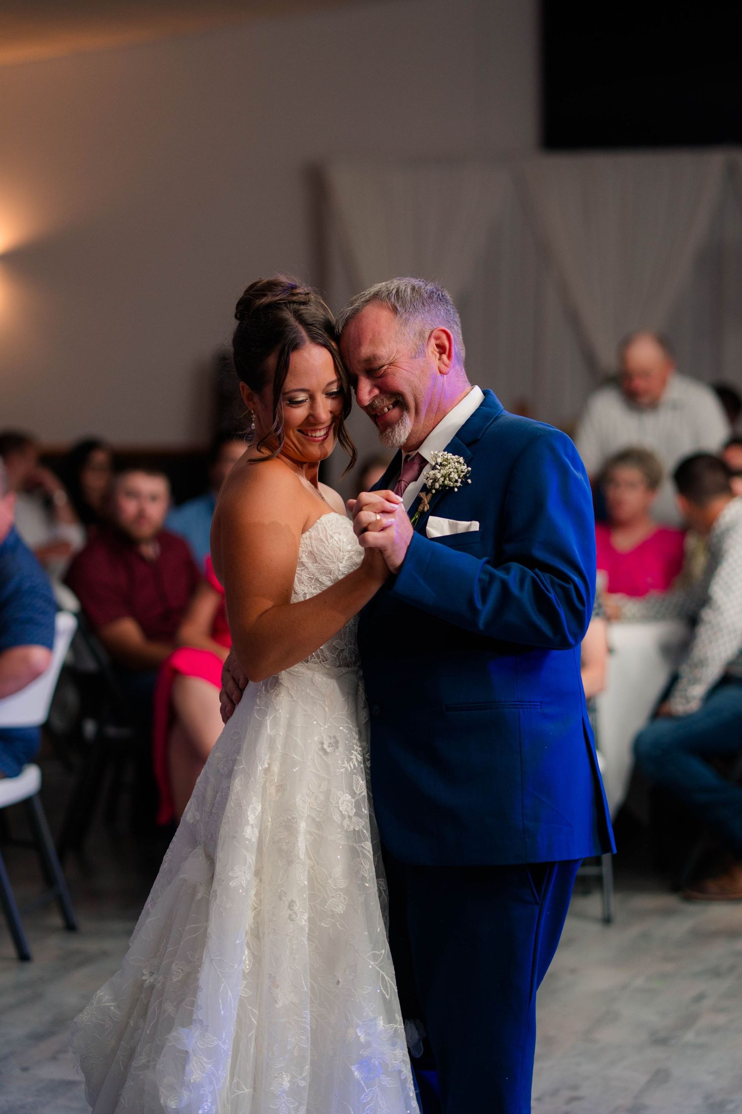 Bride and groom share their first dance, smiling joyfully, with wedding guests seated in the background.
