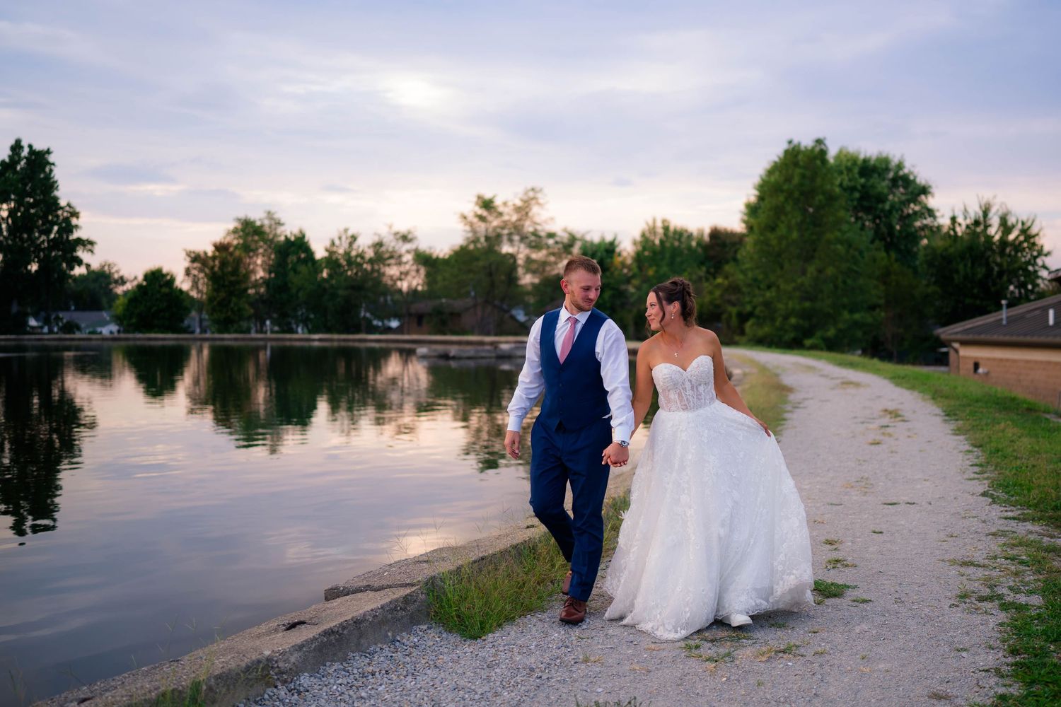 Bride and groom walk hand-in-hand along a scenic lakeside path at sunset, surrounded by lush green trees.