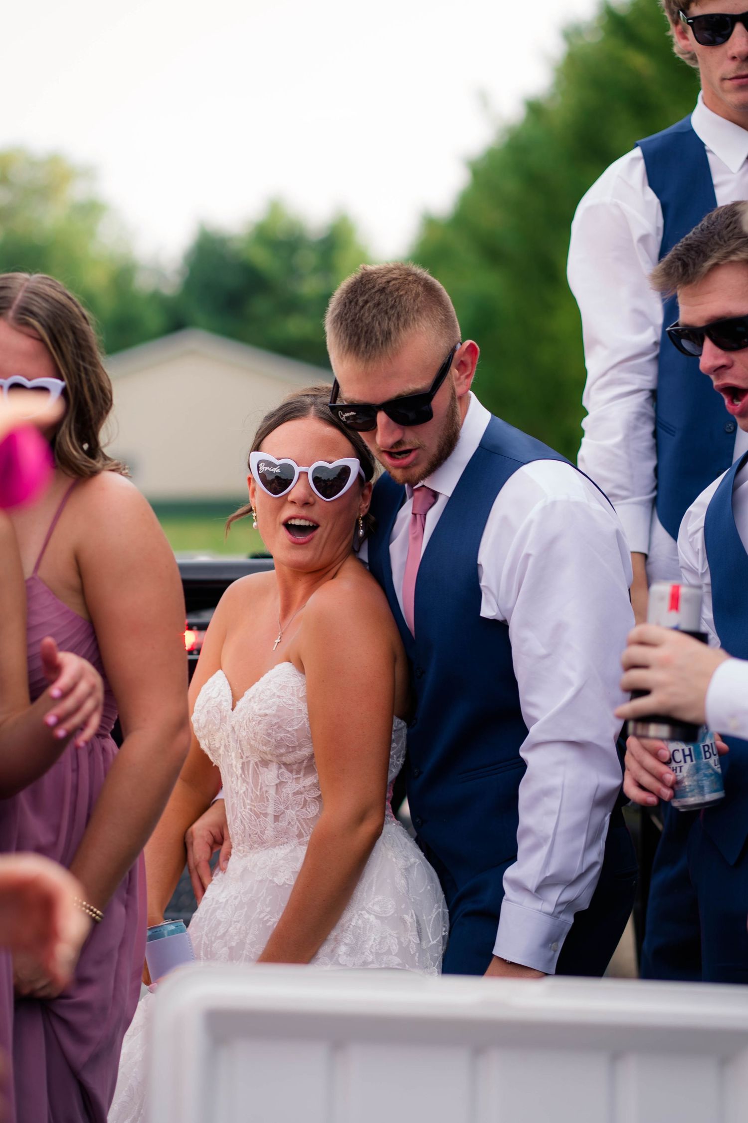 Bride and groom wearing sunglasses laugh together with wedding party during fun outdoor reception celebration.