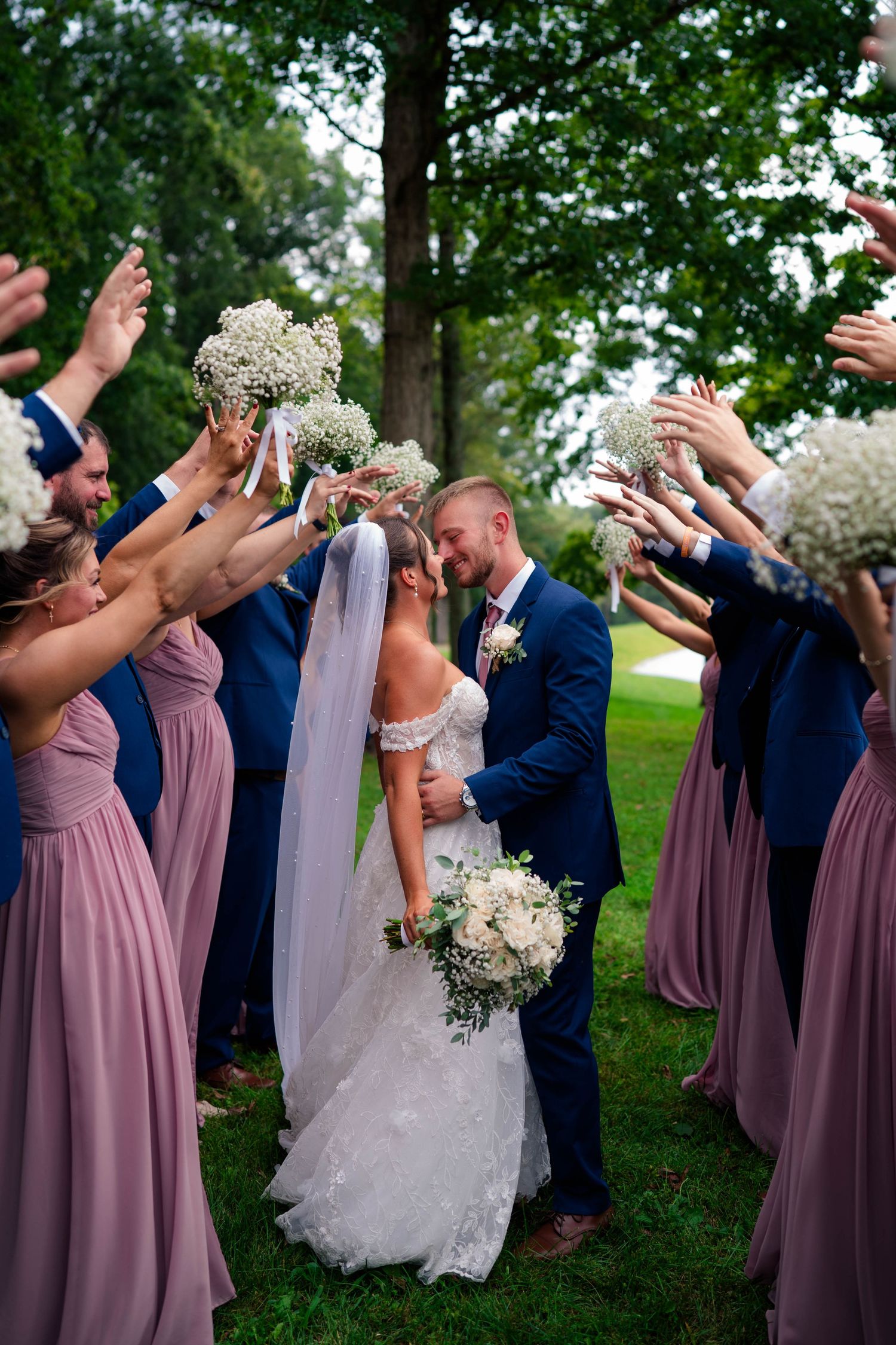 Bride and groom kiss as wedding party raises baby's breath bouquets in a celebratory arch outdoors.
