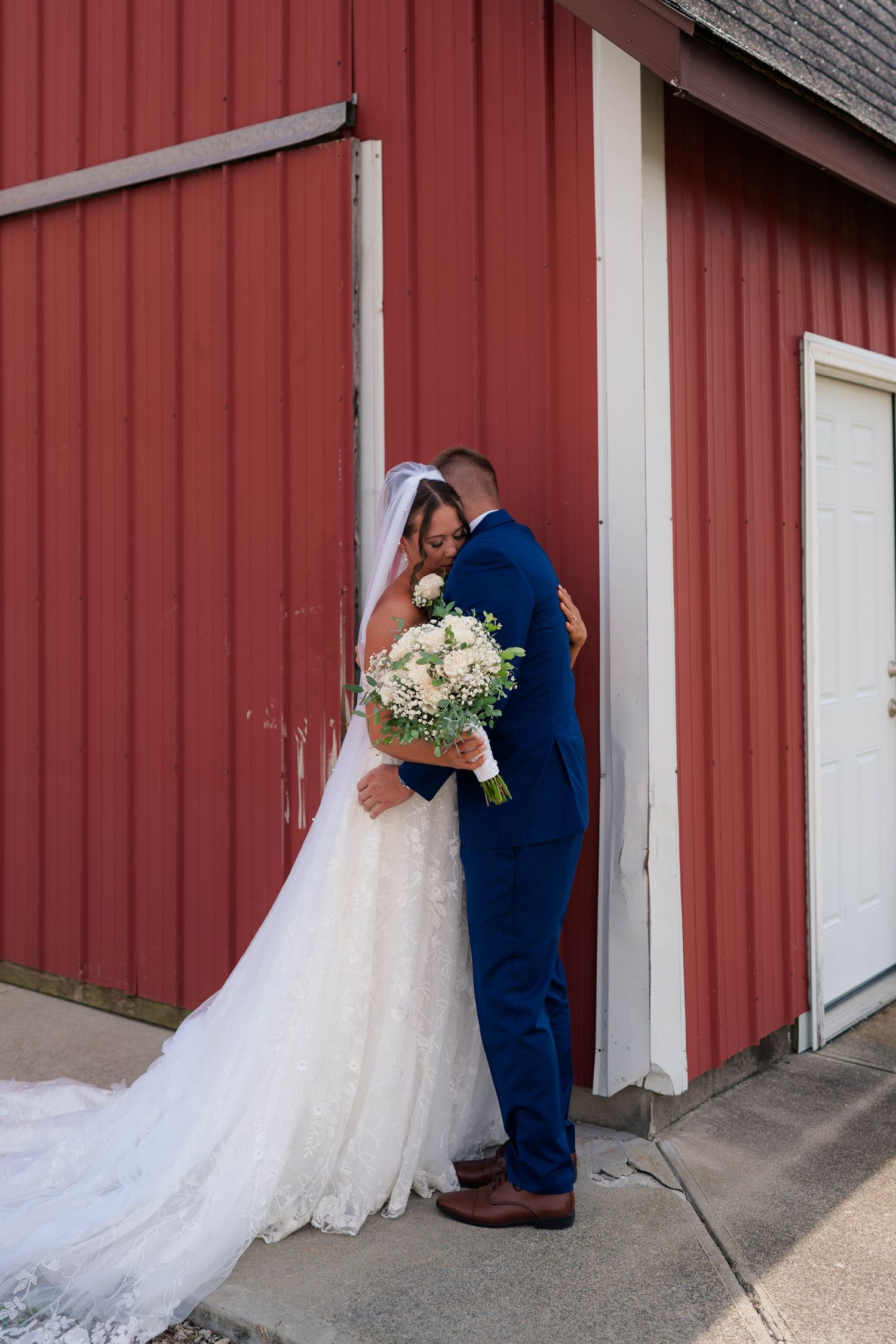 Bride and groom share an intimate embrace against a red barn wall, bride holding a white bouquet in a lace gown.