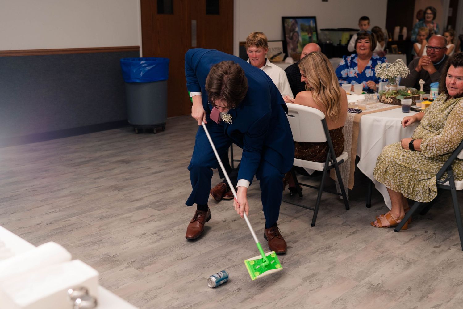 Wedding guest in navy suit playfully mops dance floor with green mop while guests watch and laugh.