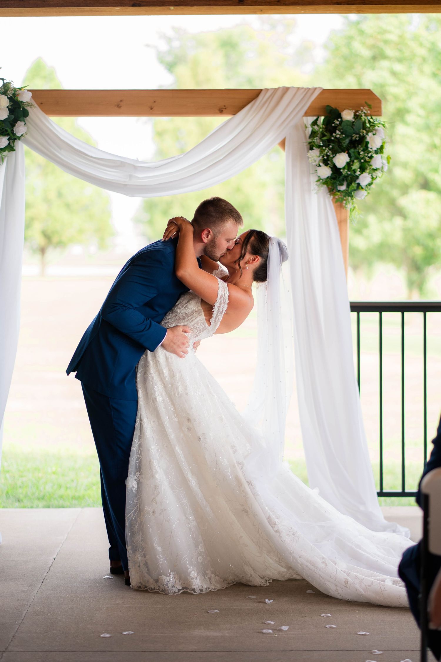Bride and groom share first kiss under wooden arch with white draping and greenery at outdoor wedding ceremony.
