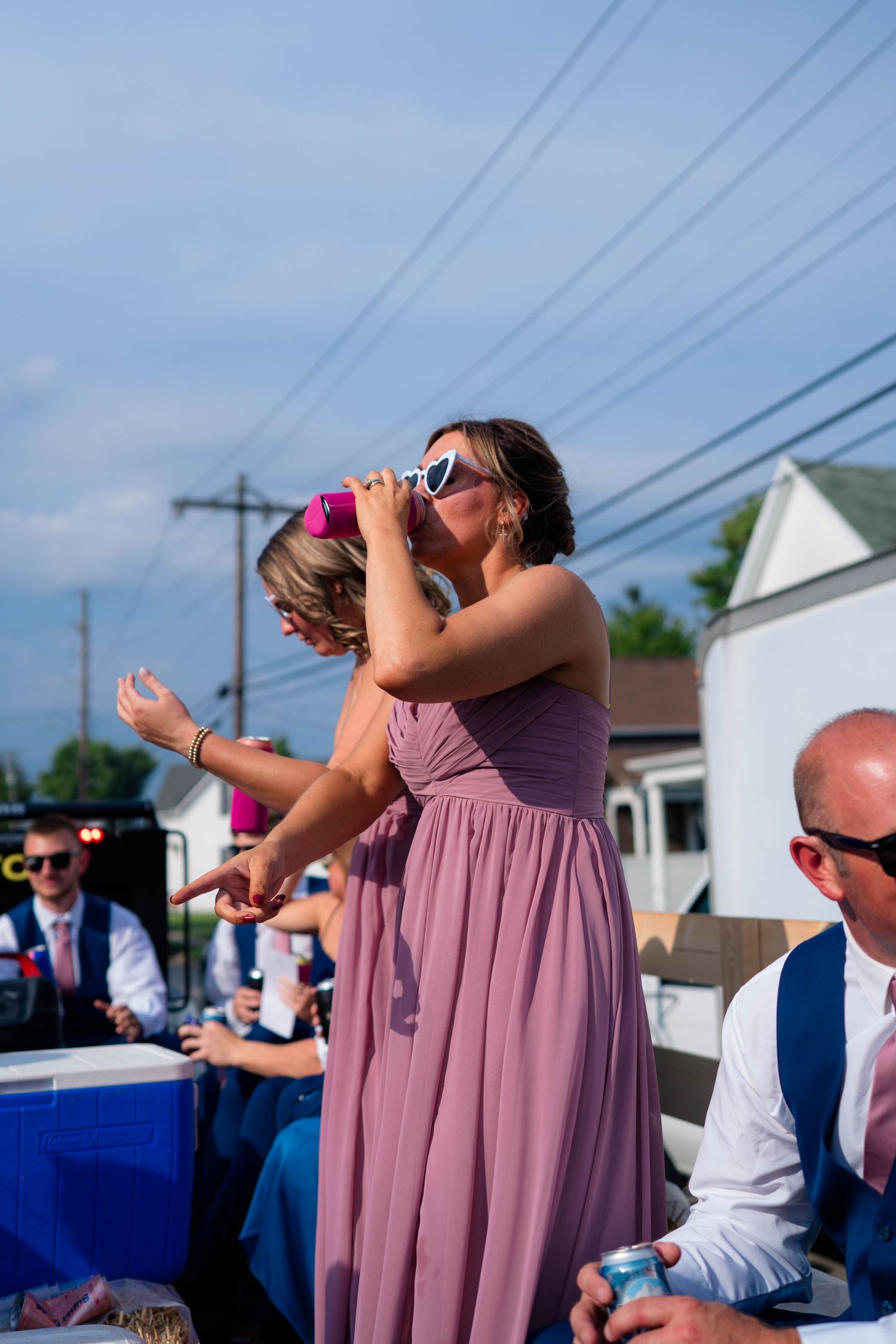 Bridesmaid in mauve dress with heart sunglasses drinking at an outdoor wedding celebration.