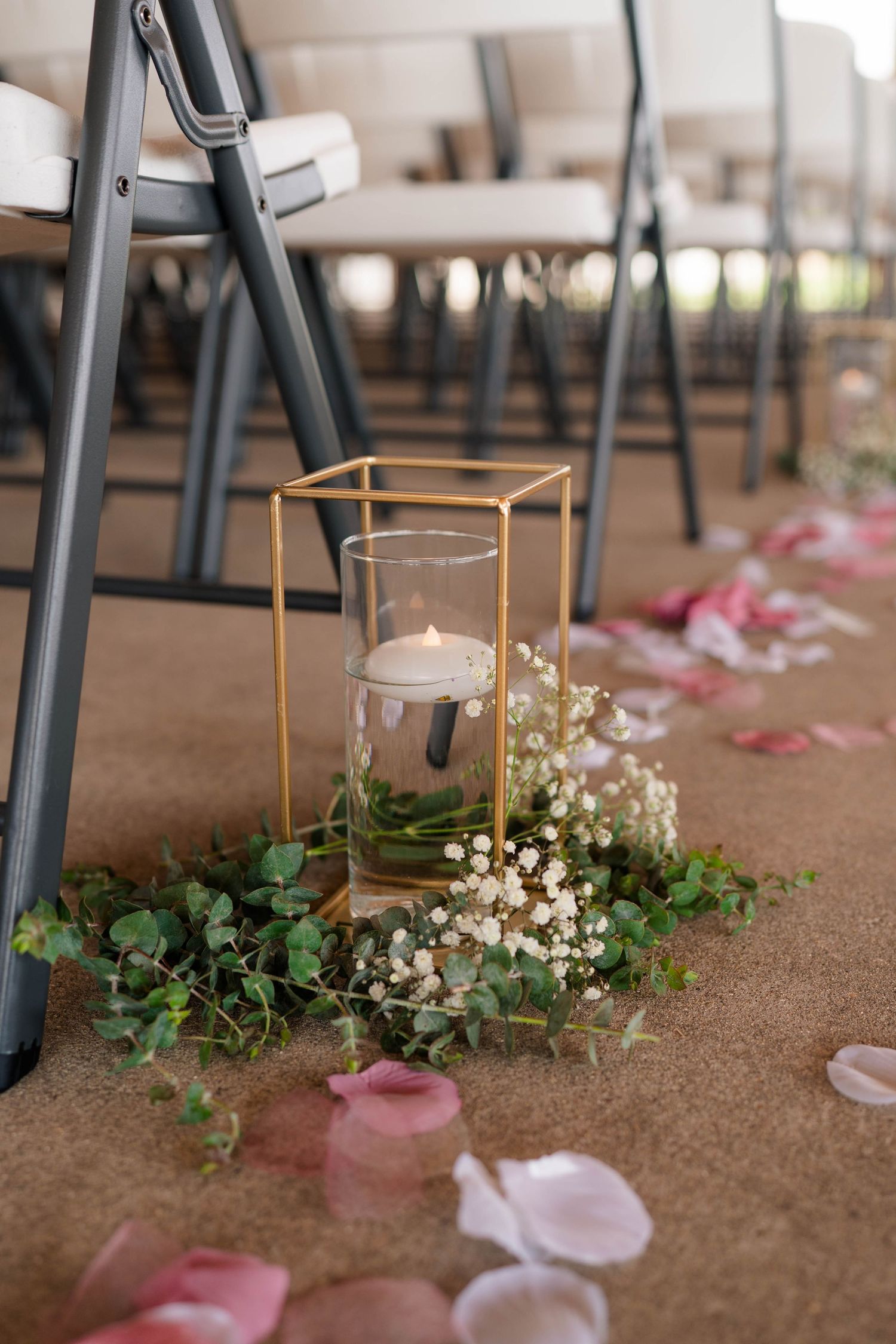Gold geometric candle holder with floating candle, greenery, and baby's breath on carpet aisle with pink rose petals at wedding.