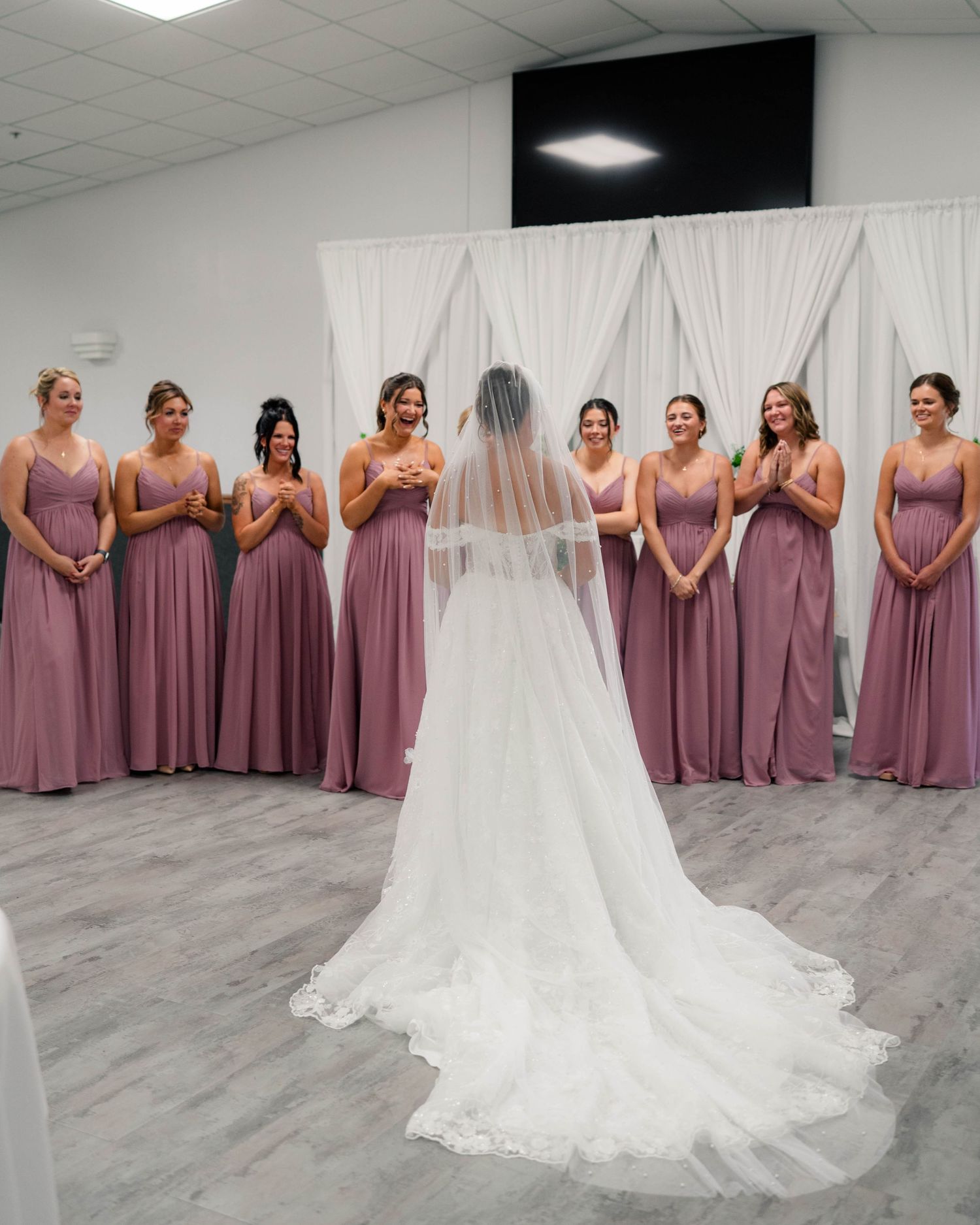 Bride in white lace gown with veil reveals herself to eight bridesmaids in dusty rose dresses.