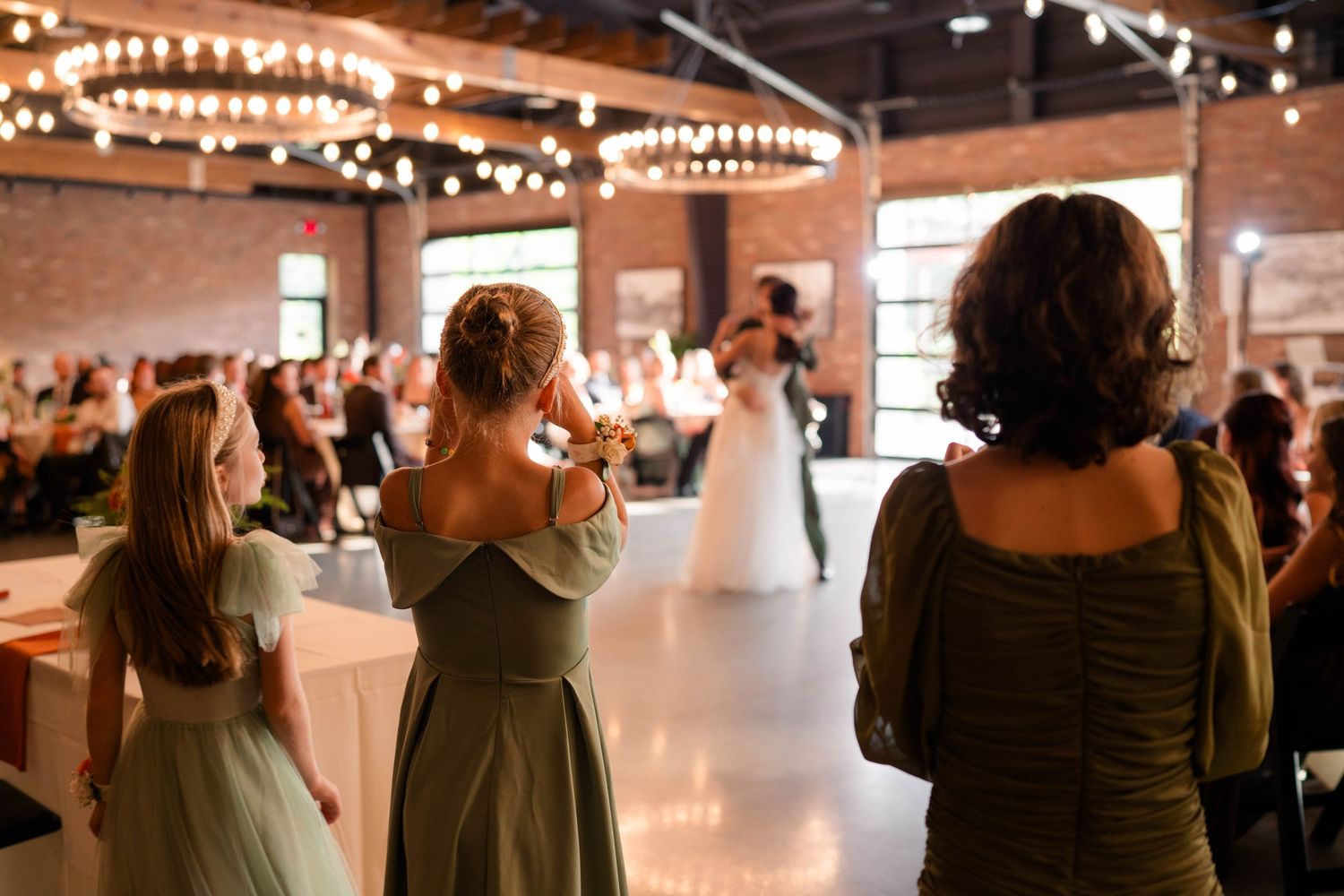 Guests in sage green dresses watch bride and groom share their first dance at a rustic reception venue.