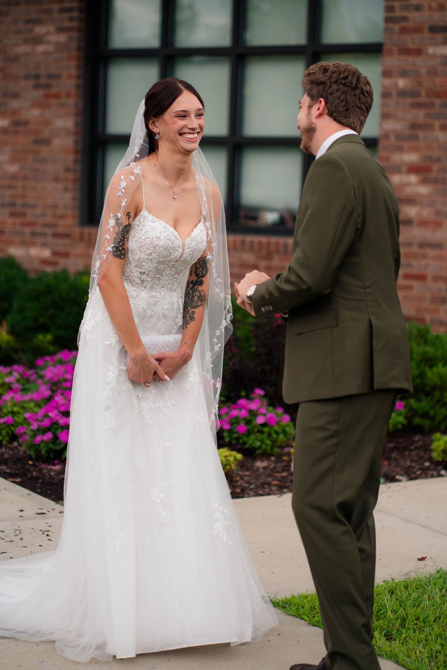 Joyful bride in lace wedding gown and veil sharing first look moment with groom in olive suit.
