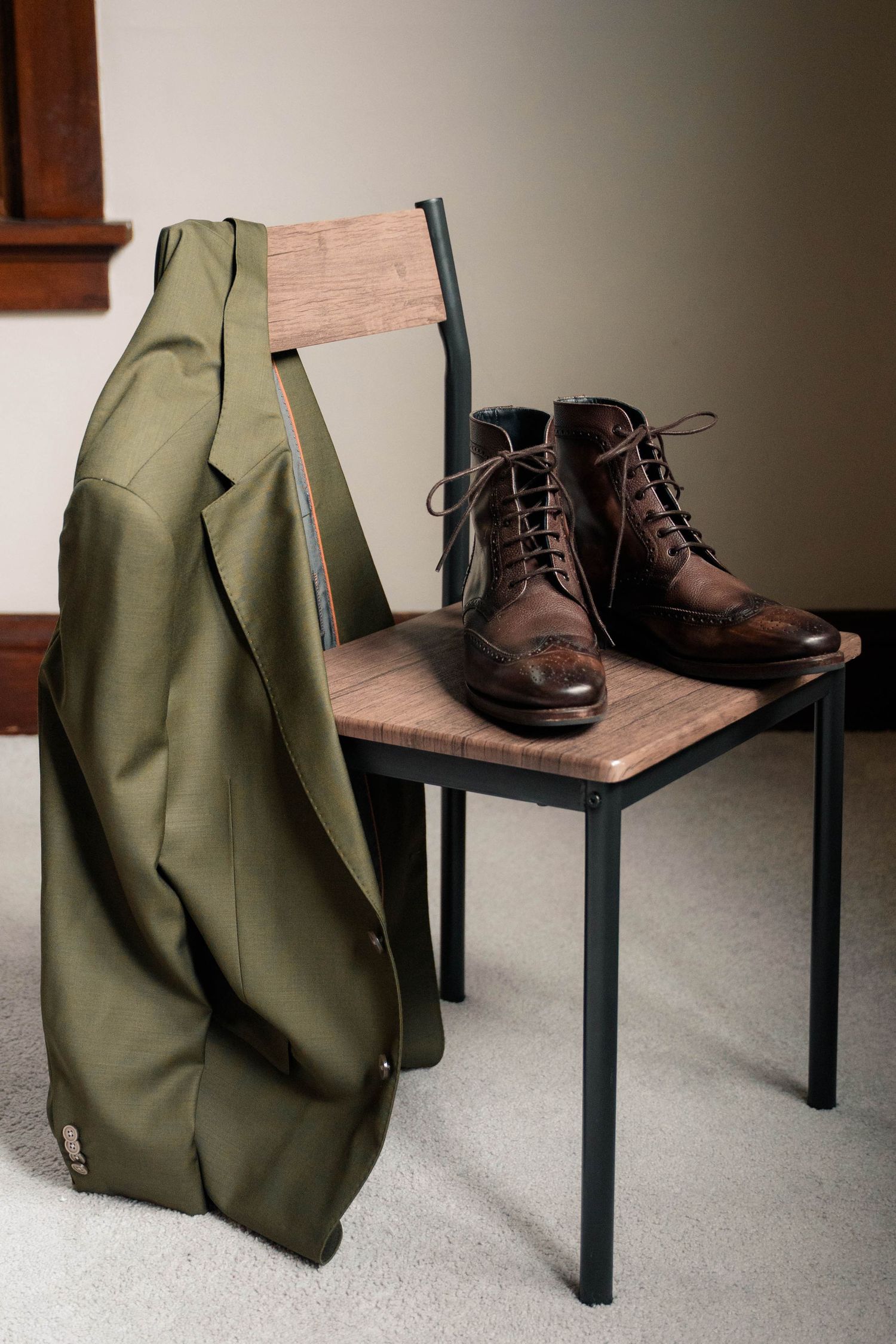 Brown leather brogue boots on a wooden chair beside a draped olive green suit jacket.