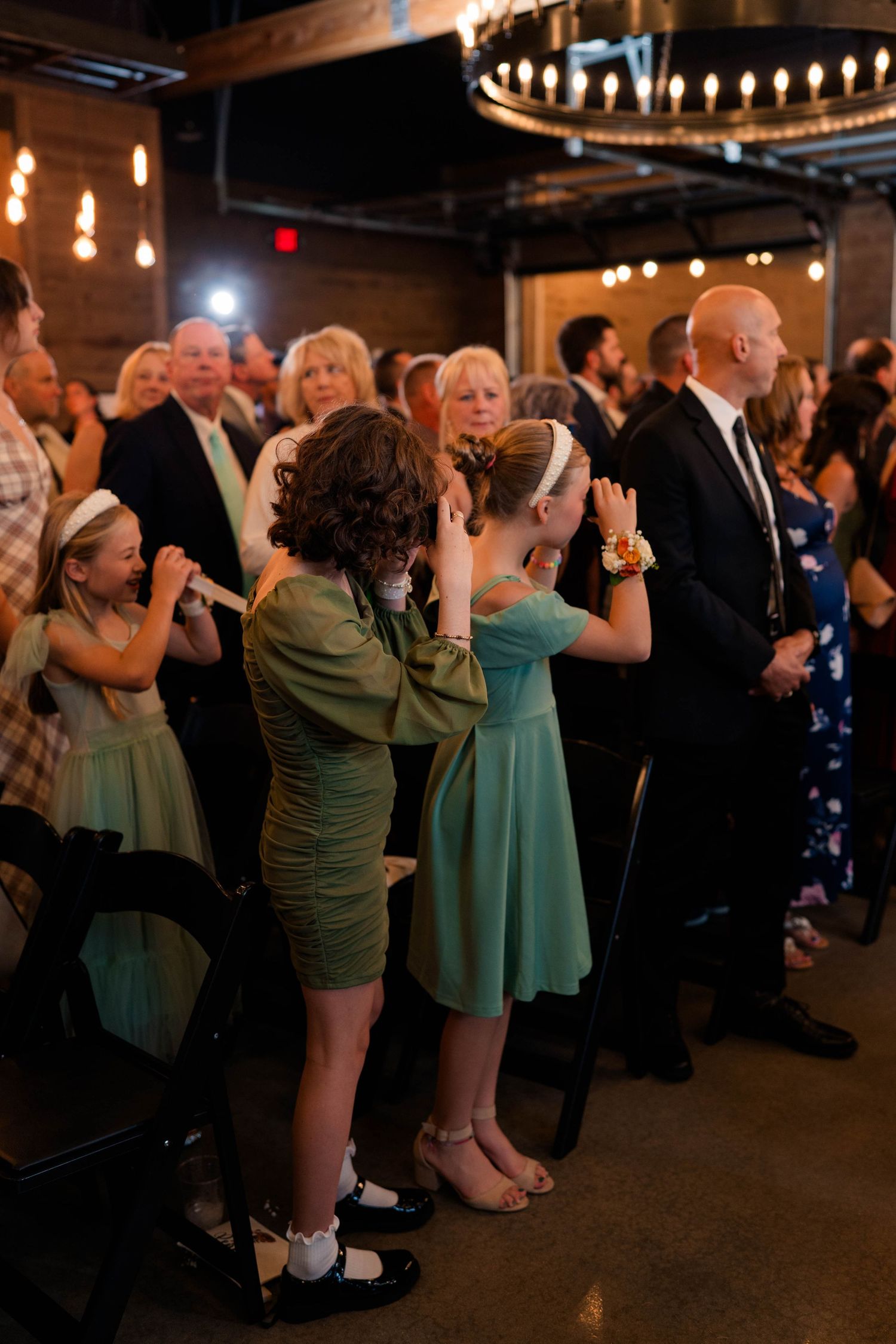 Wedding guests including young girls in mint dresses watching ceremony in rustic venue.