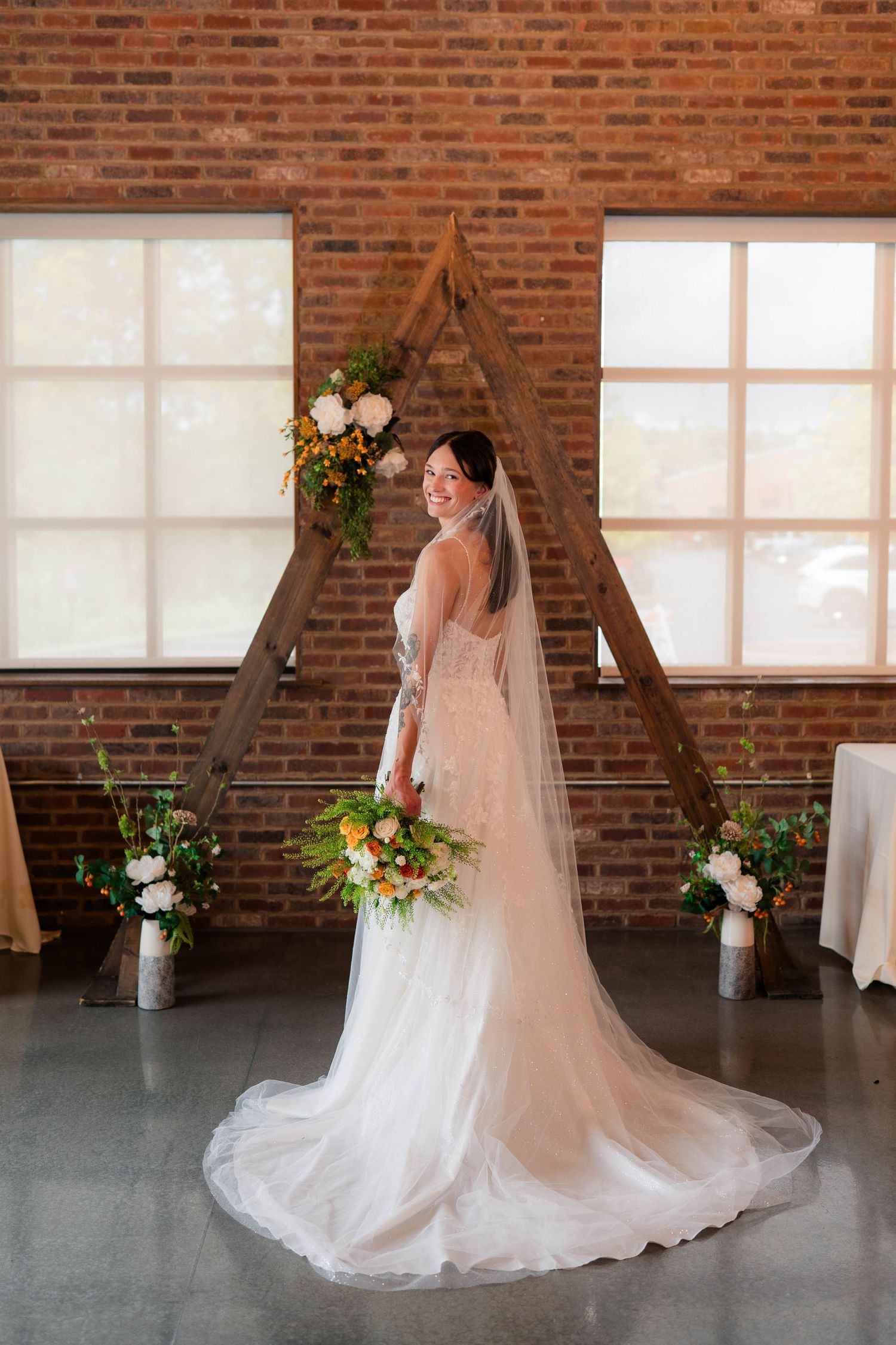 Bride in flowing lace gown with cathedral veil poses before wooden triangular arch in brick venue.