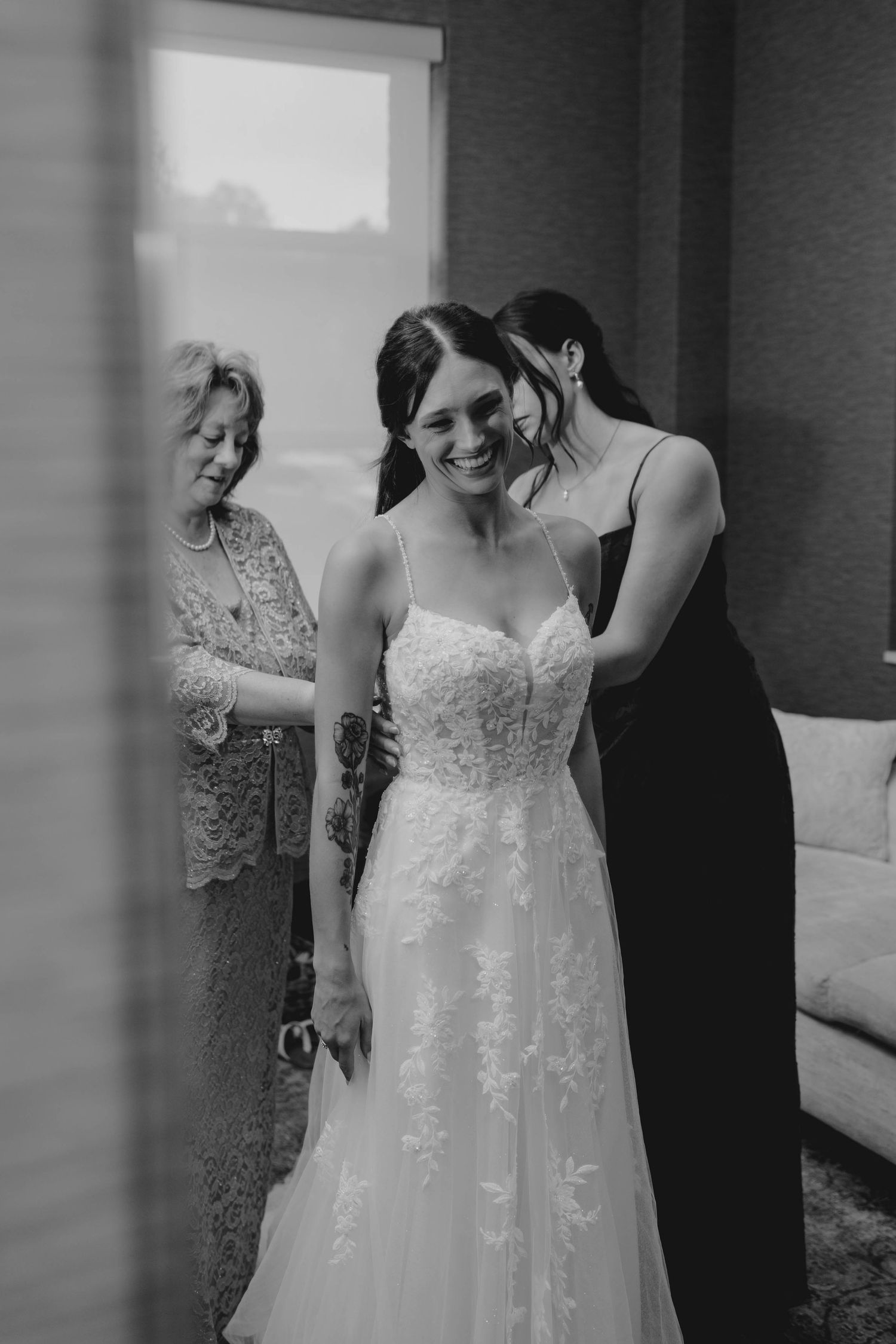 Bride smiling joyfully as bridesmaids help her into her lace wedding gown in a black and white photo.