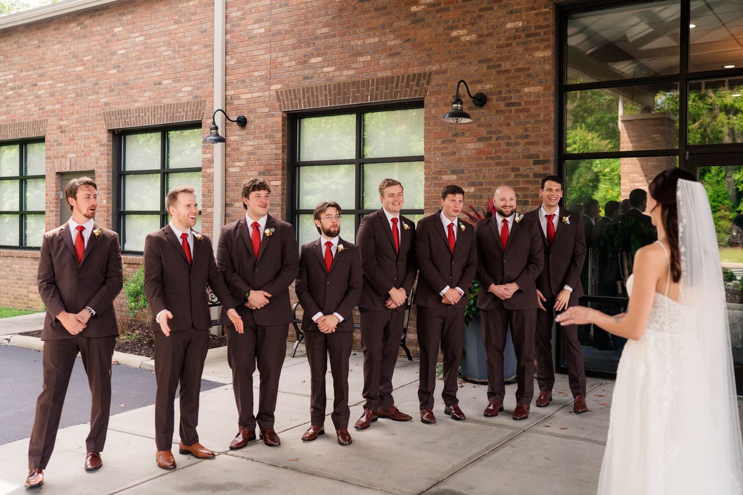 Groomsmen in brown suits with red ties reacting to bride reveal outside brick venue.
