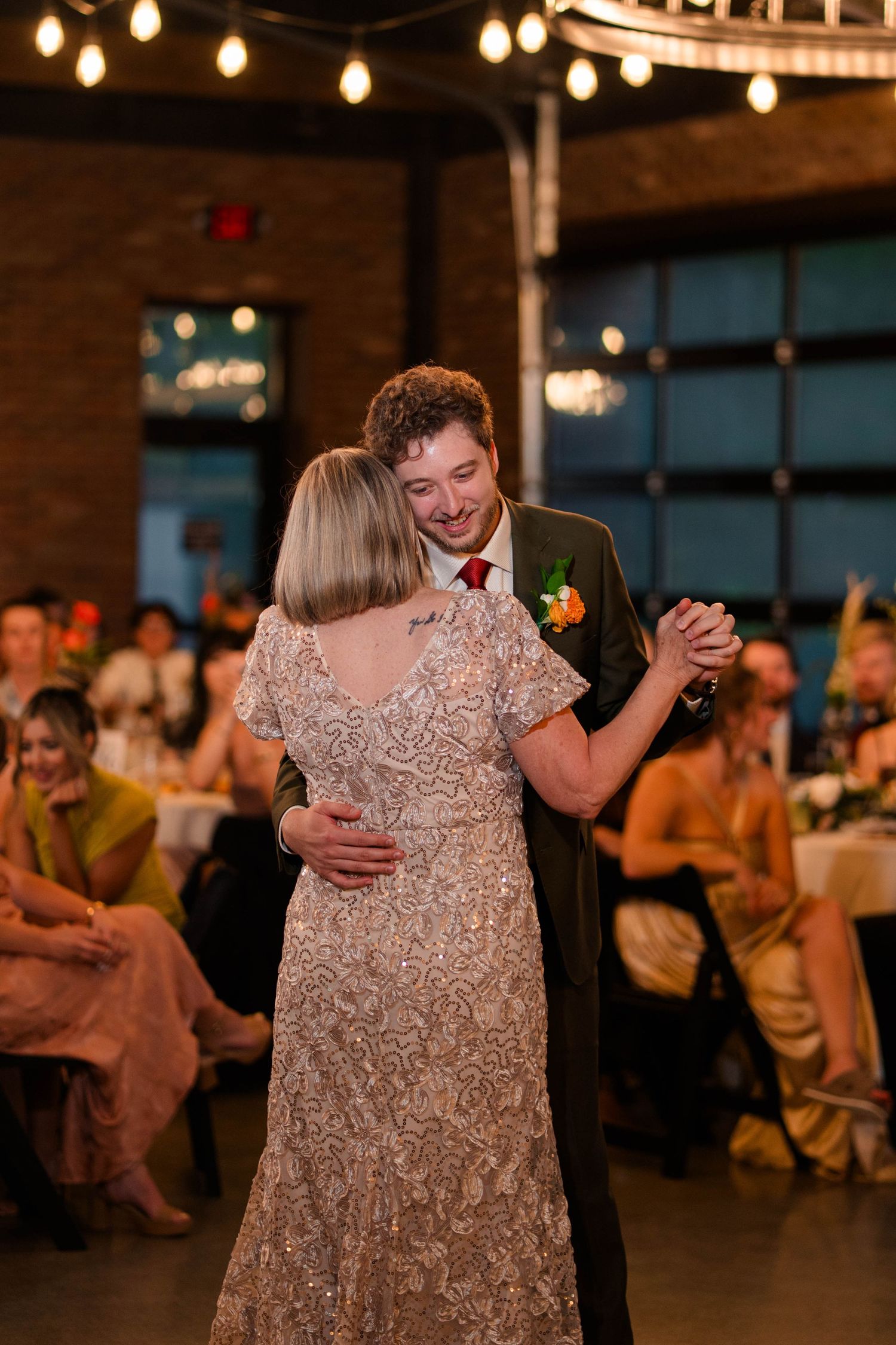 Groom smiles warmly while sharing mother-son dance at elegant wedding reception with string lights.
