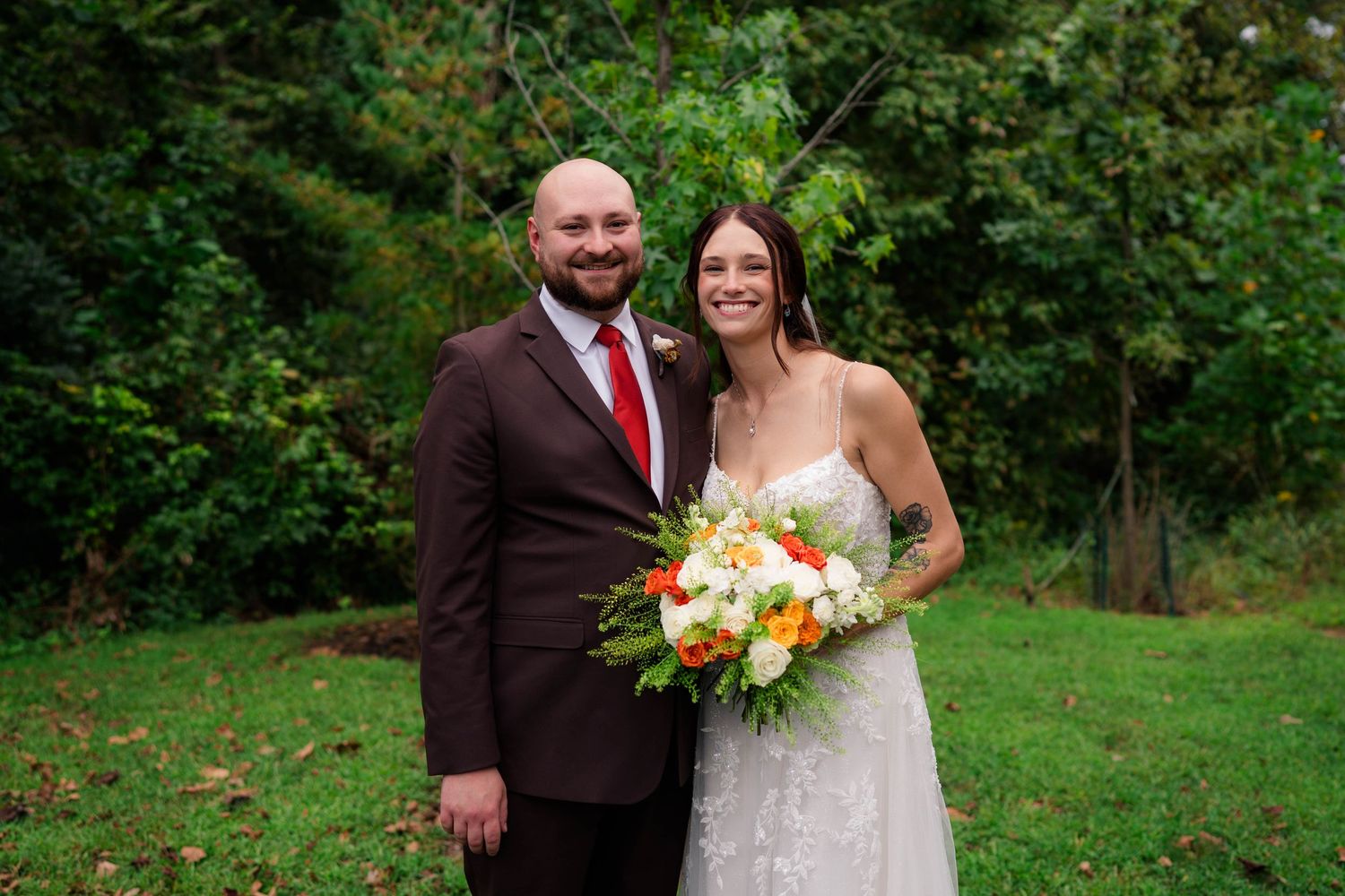 Smiling bride and groom pose outdoors, bride holding a vibrant orange and white bouquet surrounded by greenery.