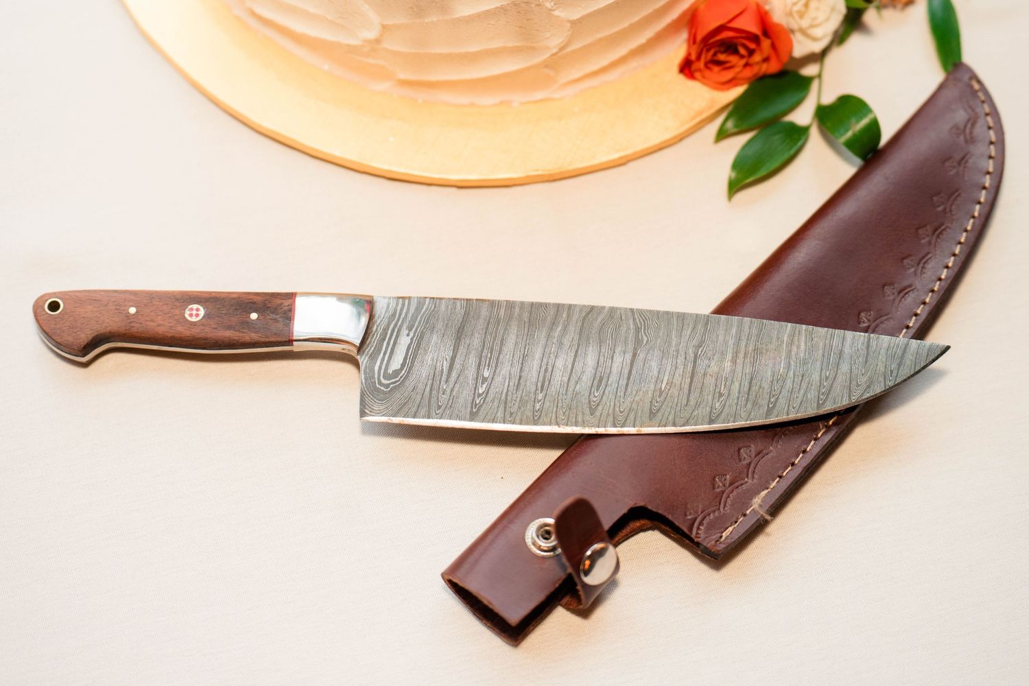 Damascus steel chef knife with leather sheath beside orange rose on white surface.