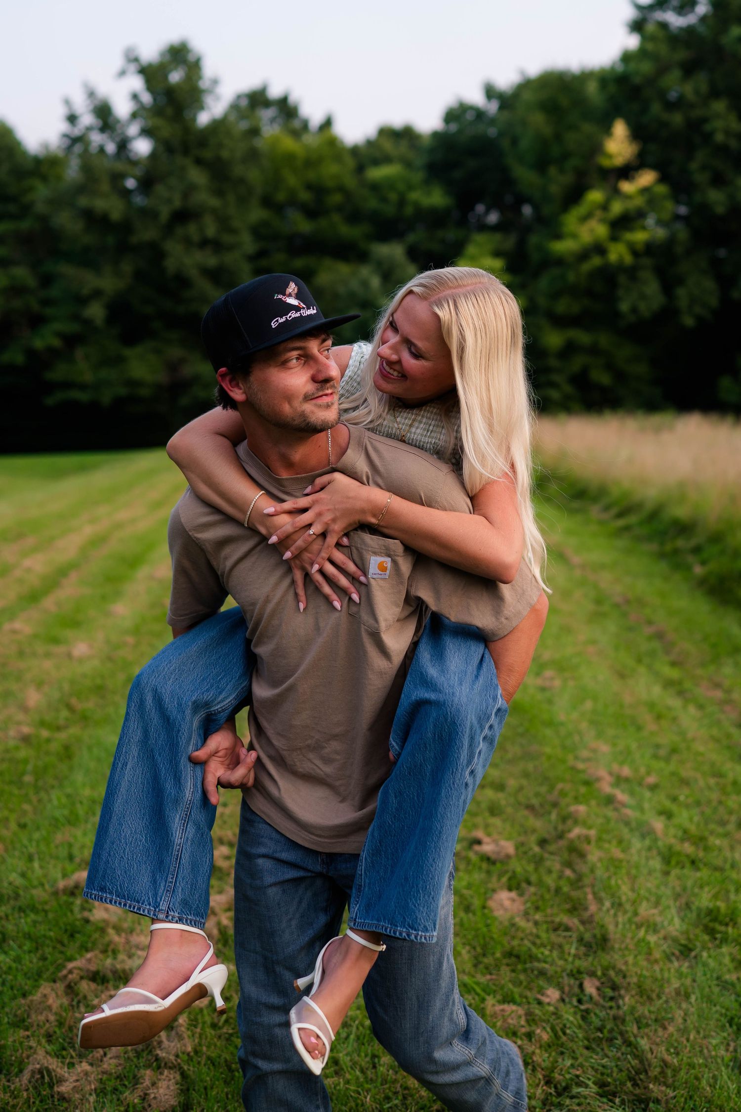 A man gives a blonde woman a piggyback ride in a green field, both smiling and wearing jeans.