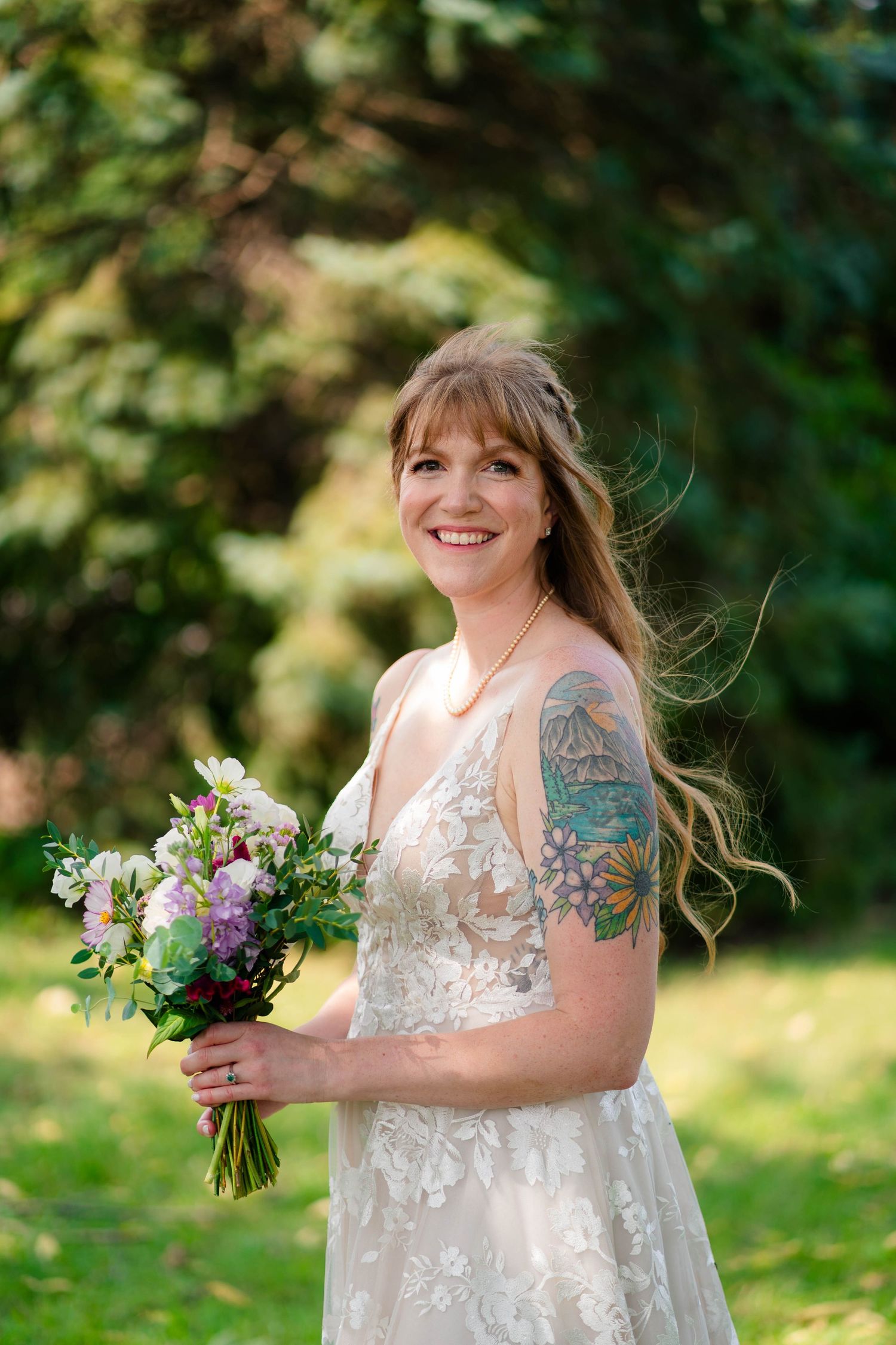 Smiling bride with tattoos holds floral bouquet in garden, wearing elegant lace wedding gown outdoors.
