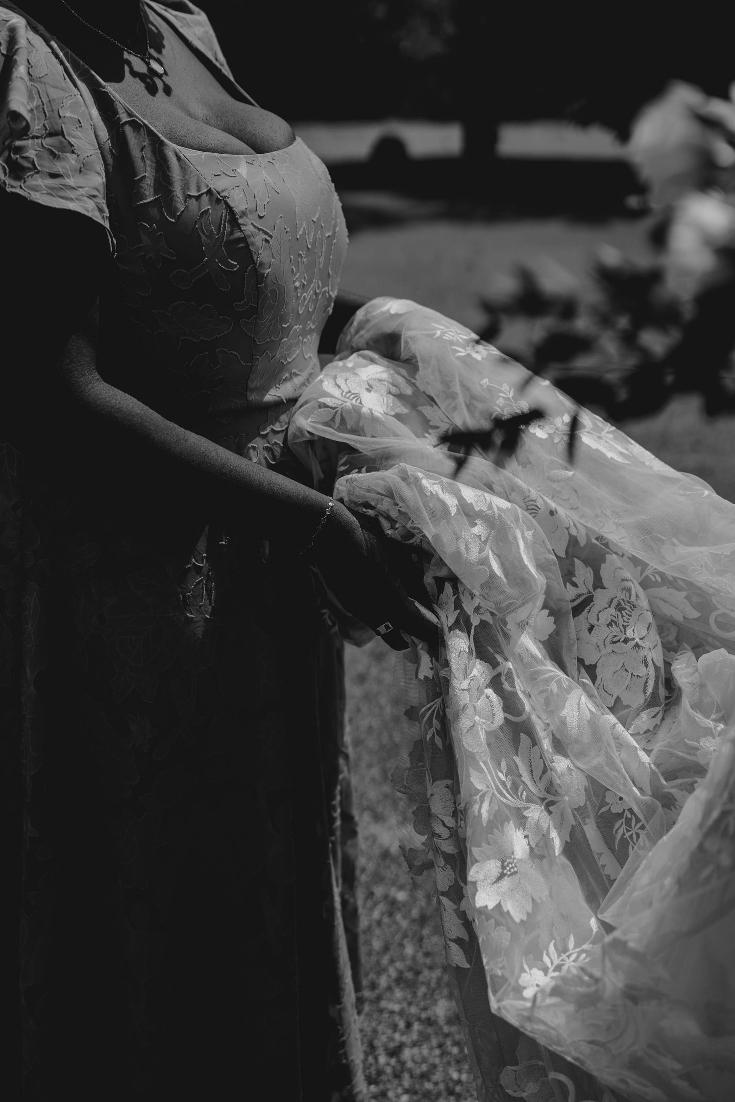 Black and white close-up of bridesmaid holding floral wedding gown train at outdoor ceremony.