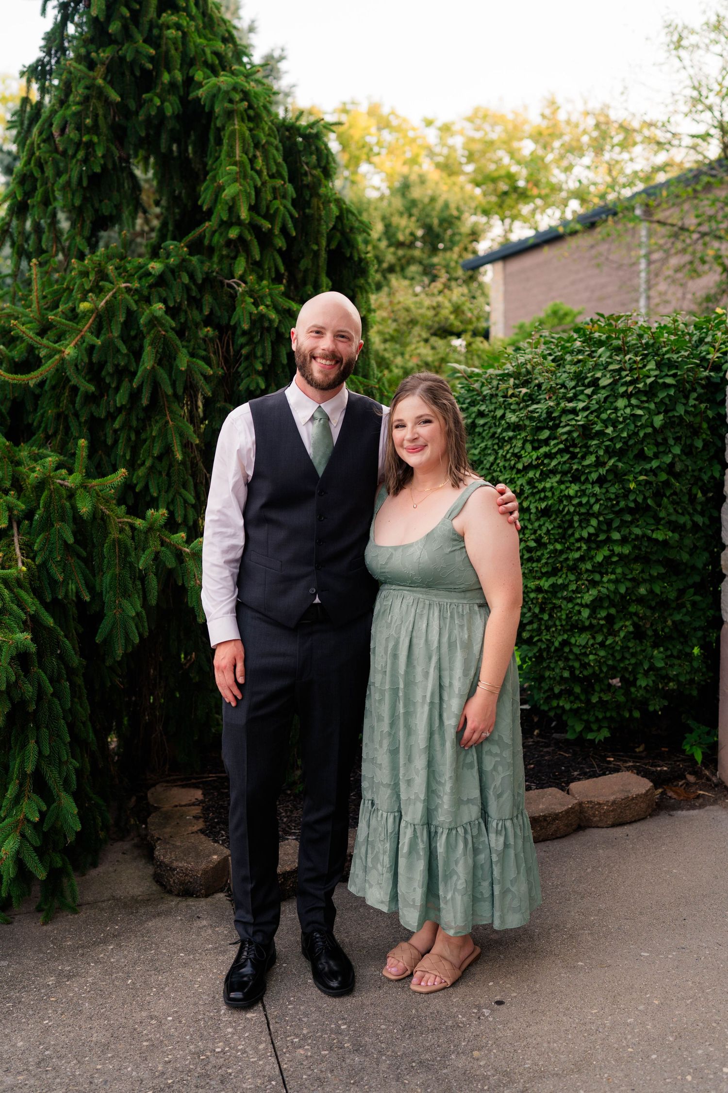 Bald groom in black vest and green tie poses outdoors with woman in sage green dress.