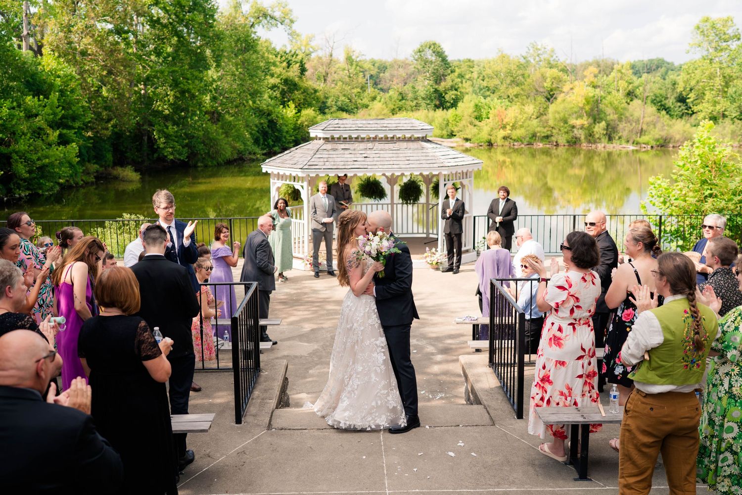Bride and groom share first kiss at outdoor lakeside wedding ceremony with guests watching from both sides.